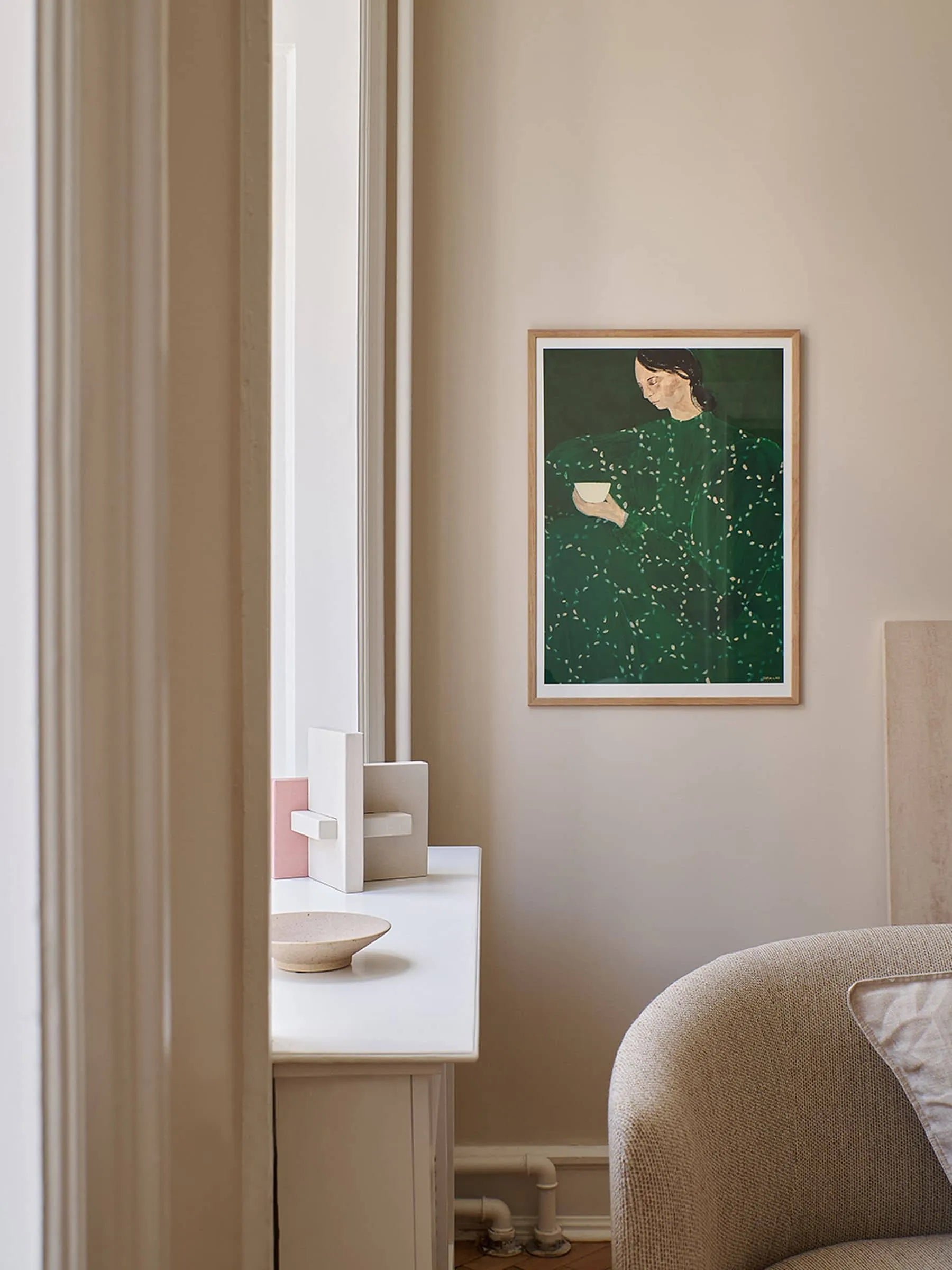 A cozy, neutral-toned room features a curved beige armchair, minimalist art on the windowsill, and The Poster Clubs “Sofia Lind - Coffee Alone At Place De Clichy (30x40)” on the wall—a perfect blend of style and fine art.