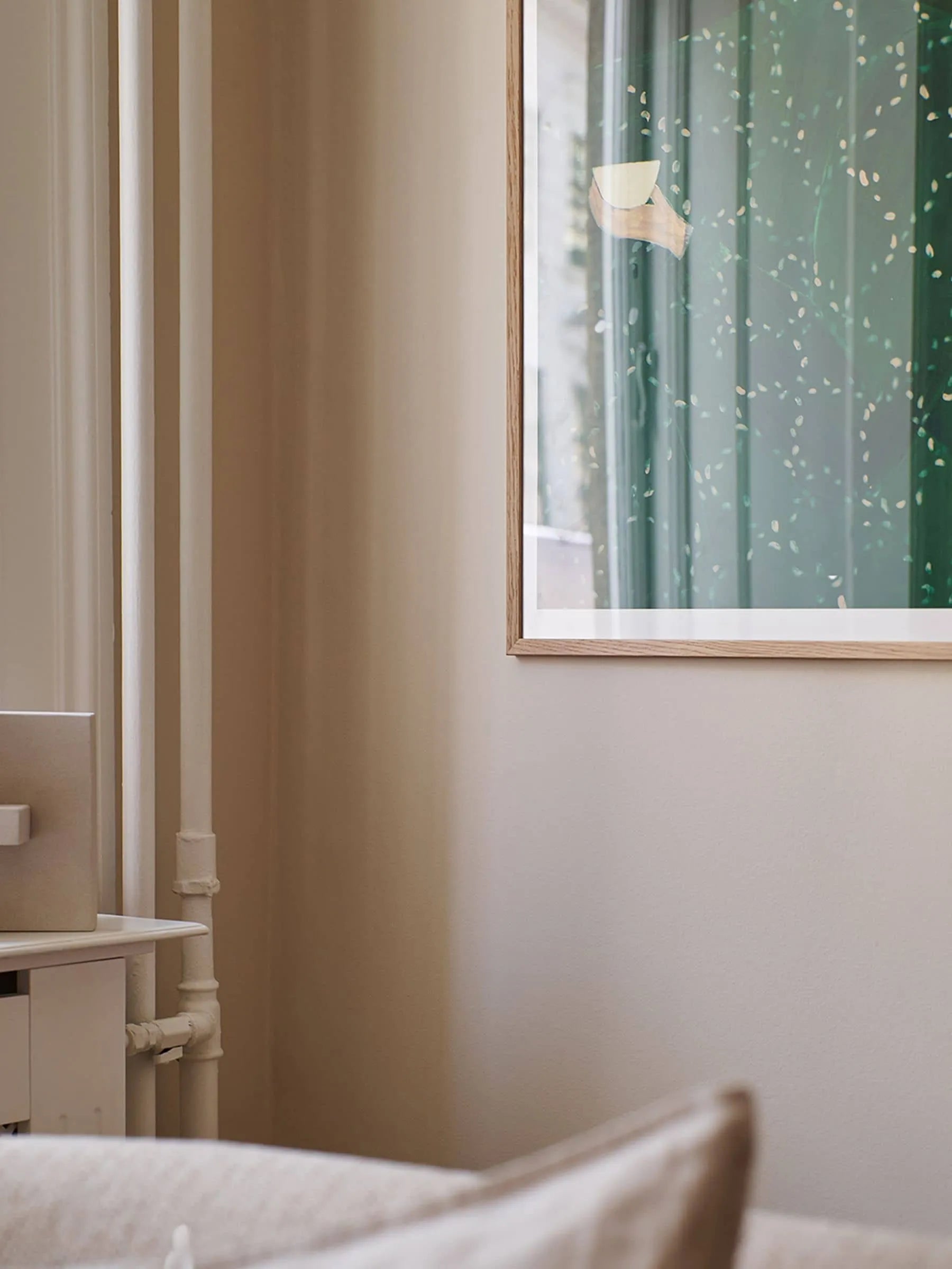A softly lit, minimal corner with beige walls features part of The Poster Clubs Sofia Lind - Coffee Alone At Place De Clichy (30x40) giclée print, showing a green abstract design beside a white cabinet and exposed pipes.