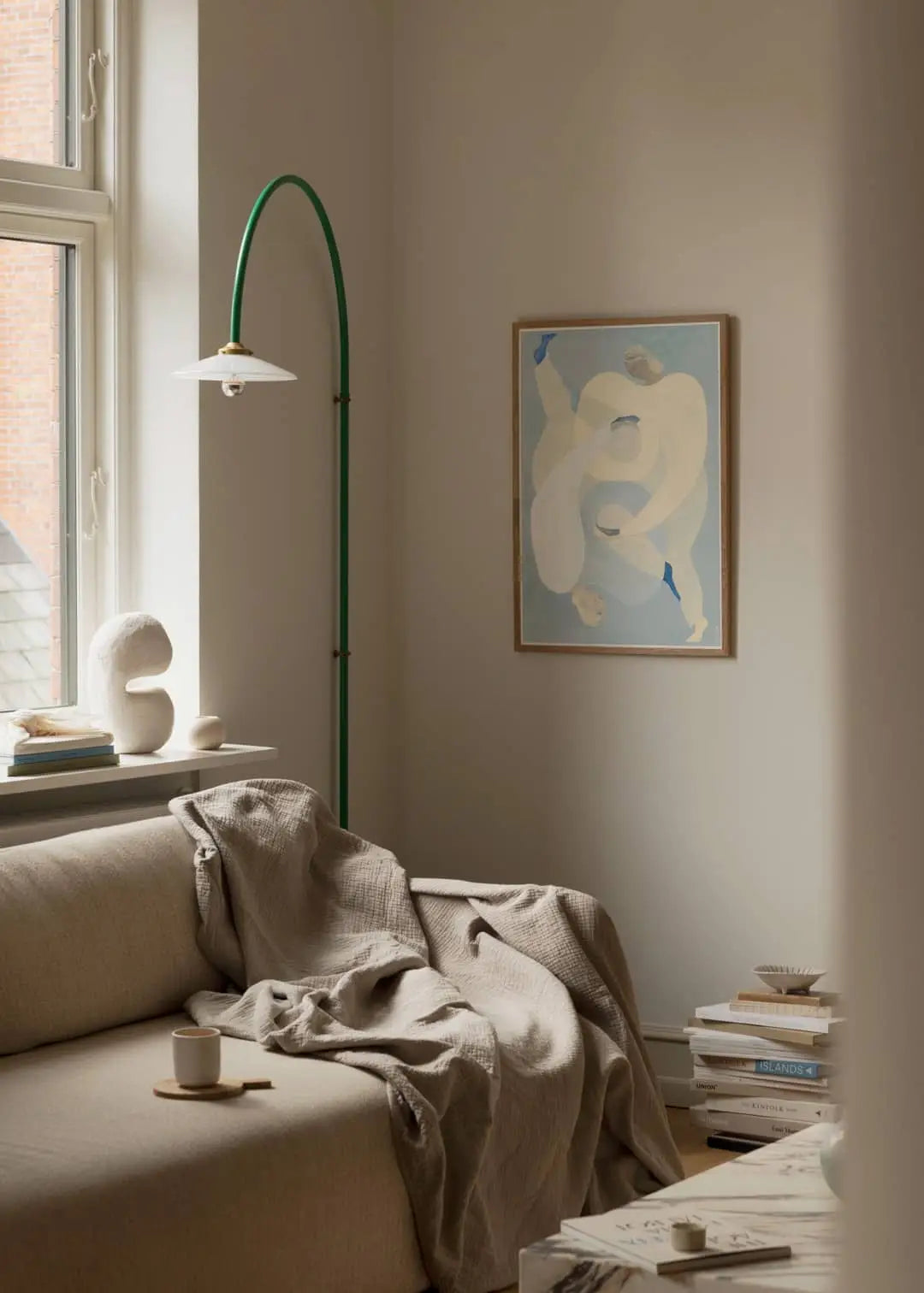 A cozy living room with a beige sofa, textured blanket, cup on coaster, books, and modern green floor lamp. Above hangs The Poster Club Berit Mogensen Lopez - Improvise (50x70) art print as sunlight streams in.