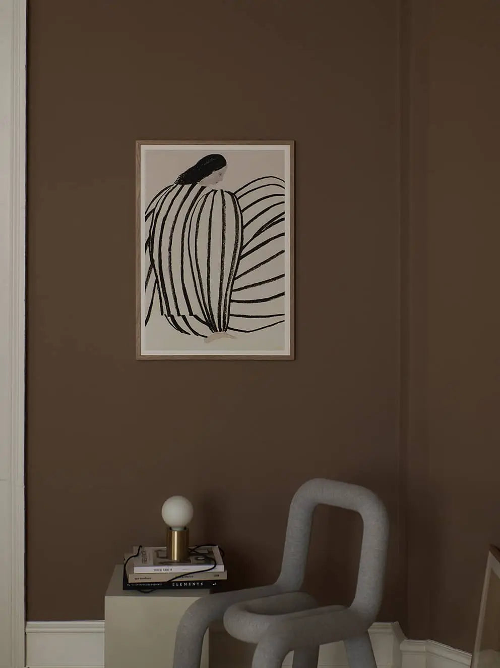 A minimal, modern room with brown walls features The Poster Club Sofia Lind - You (50x70) art in a light frame, a sculptural gray chair, a book-topped small table, and a round white lamp with a gold base.