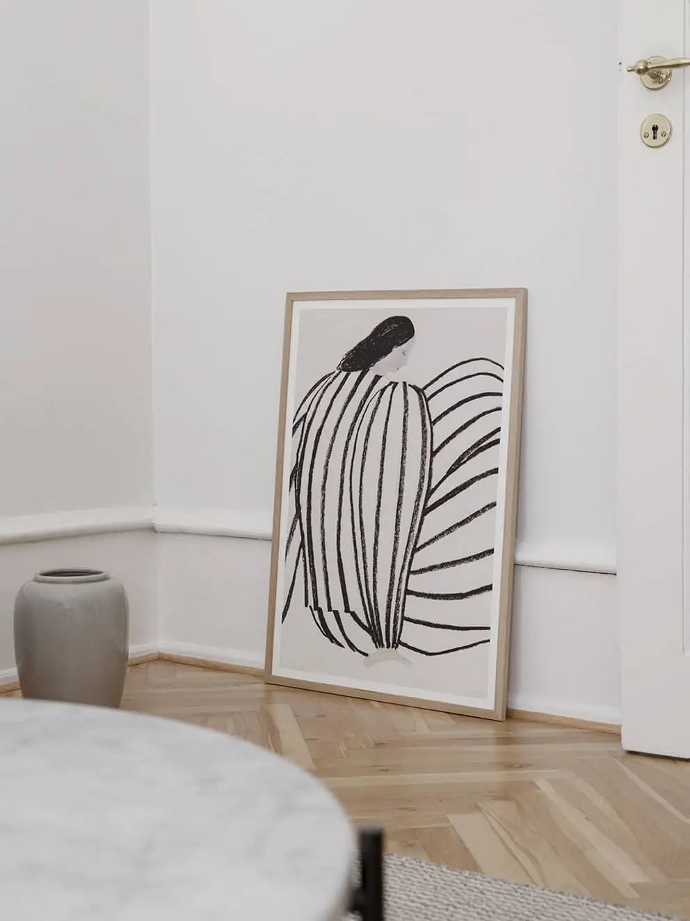 The Poster Club’s Berit Mogensen Lopez - Bloom (50x70) framed art print leans against a white wall, with a gray vase beside it on the floor and part of a round marble table from The Poster Club showing in the foreground.