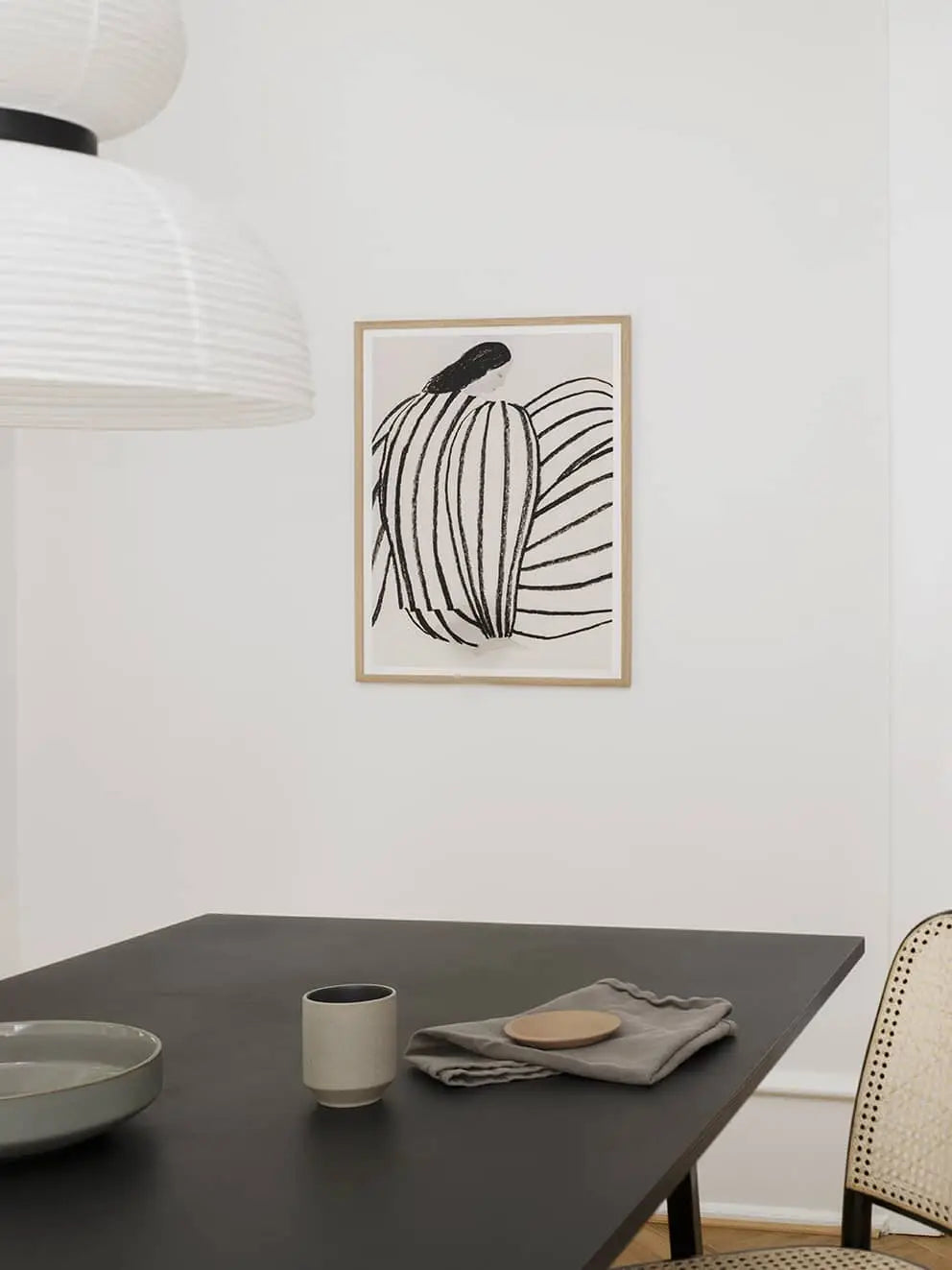 A minimalist dining area with a black table, gray mug, bowl, plate, folded napkin, rattan chair, and The Poster Club Berit Mogensen Lopez - Improvise (50x70) art print on the white wall. A white pendant light hangs above.