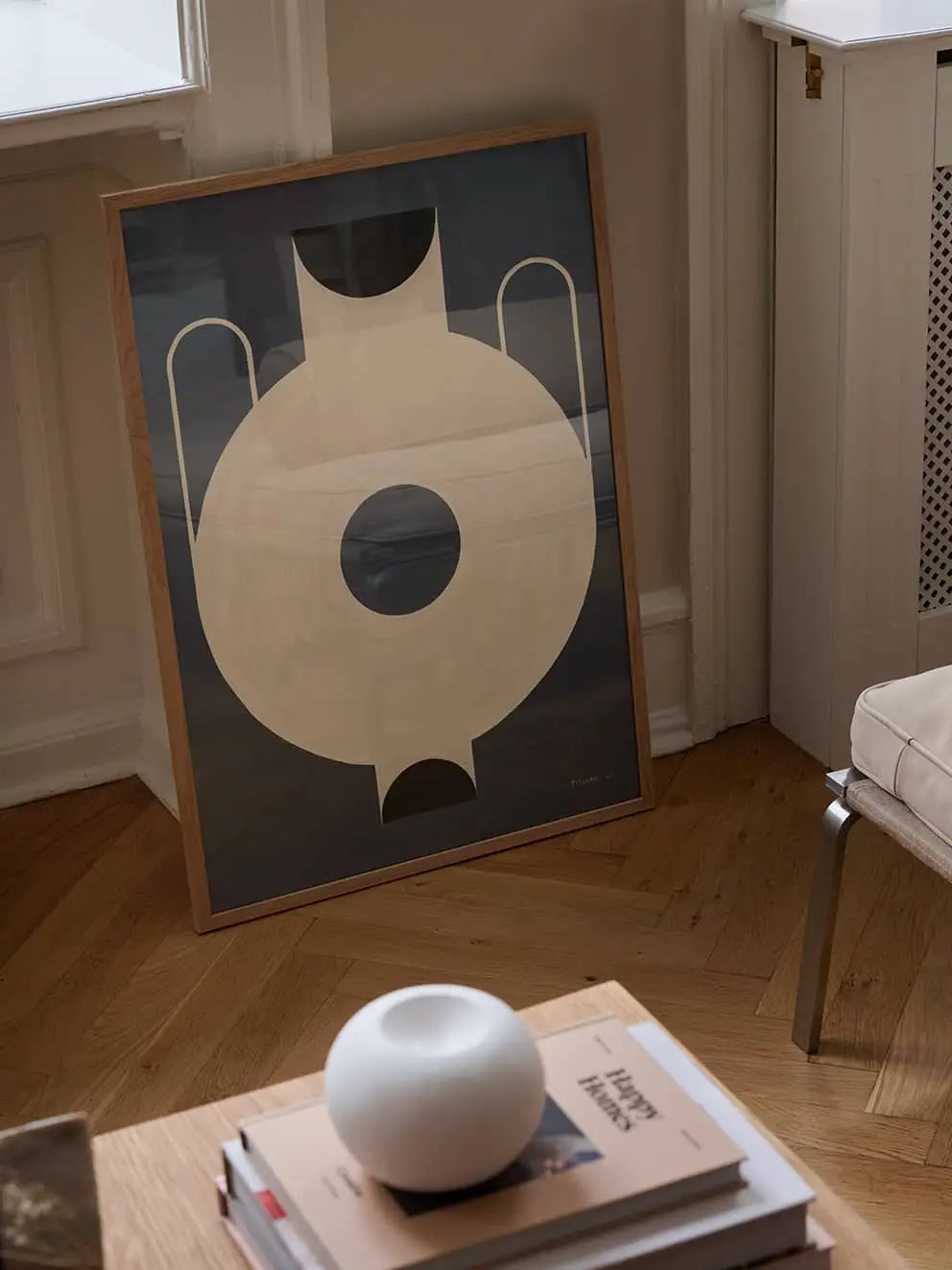 The Poster Club Studio Paradissi - Circular Vase (50x70) giclée print, with cream and black geometric shapes, leans against a wall on a wooden floor next to a chair, coffee table with books, and a round white ceramic object.