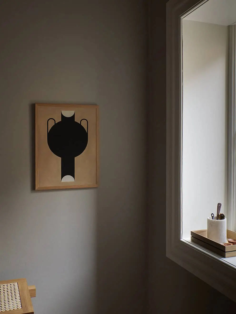 A minimal room with neutral walls features The Poster Club’s Studio Paradissi - Silhouette Of A Vase 07 (50x70) giclée print and a sunlit window above a tray with utensils in a cup and a closed book.