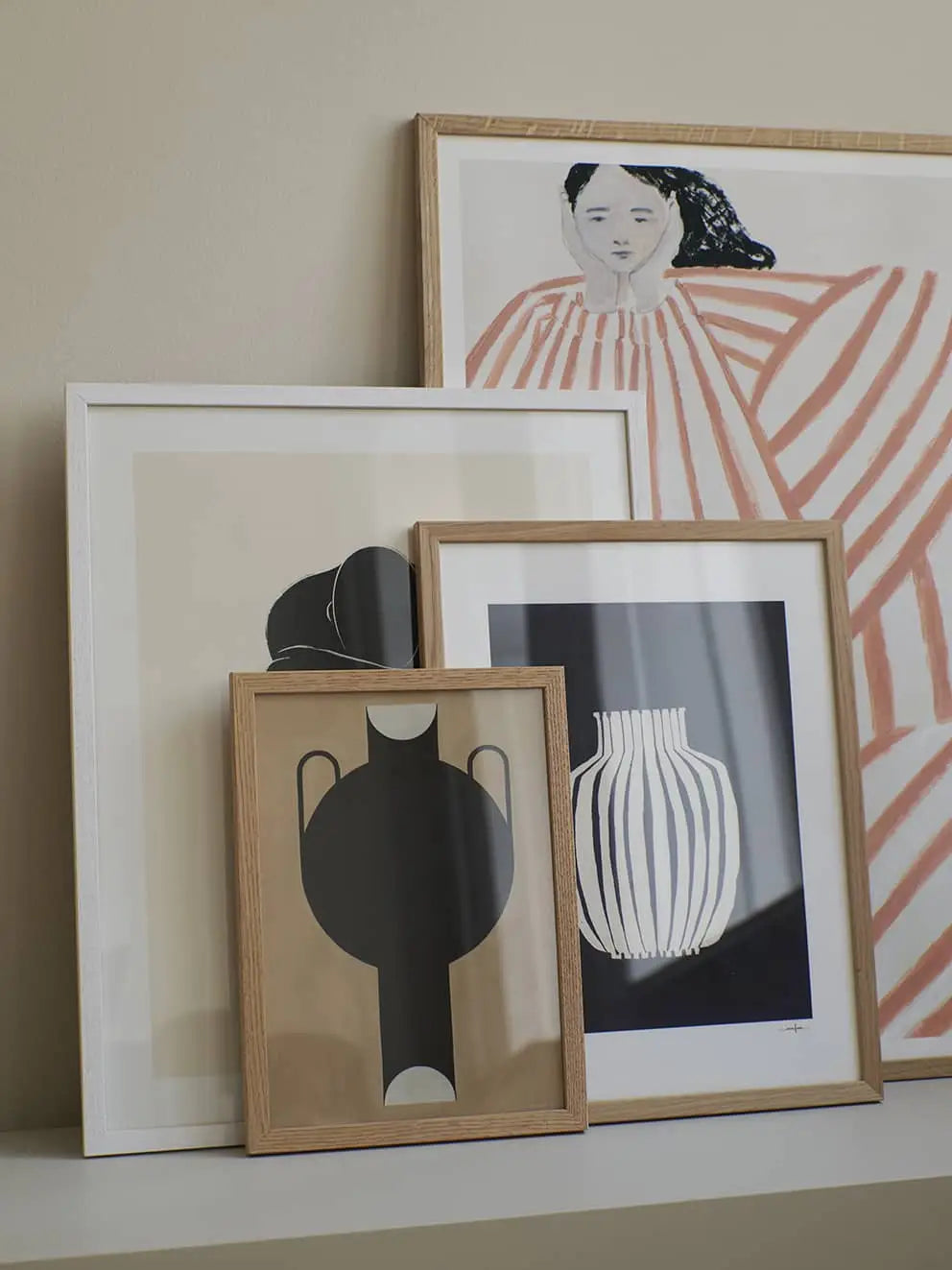 Four framed prints, including The Poster Club’s Studio Paradissi - Silhouette Of A Vase 07 (50x70), and a portrait of a woman in red and white stripes, are displayed on a white shelf against a beige wall. Frame sizes and wood tones vary.