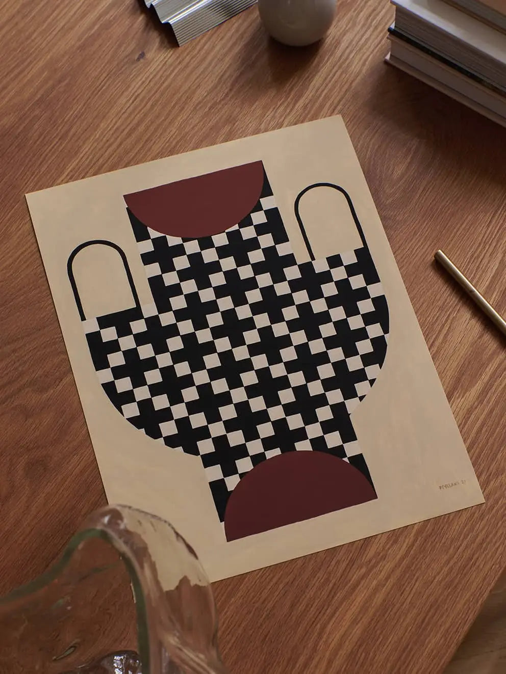 The Poster Club Studio Paradissi - Vase With Cross Pattern (50x70) giclée print by The Poster Club, displayed on a wooden table with books, a gold pen, a vase, and a small round object nearby.