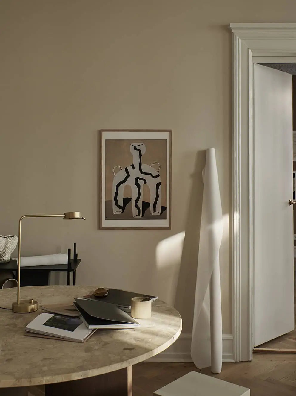 A beige room features a round marble table with books, gold desk lamp, and cup. On the wall hangs The Poster Club Studio Paradissi - Veins 01 (50x70) giclée print; tall white papers and an open door are nearby.