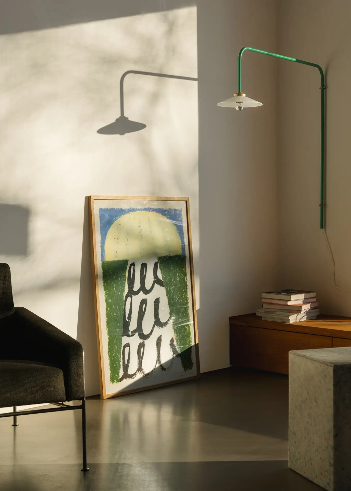 Sunlight fills a minimalist room with a modern black chair, The Poster Club Suzanne Lustig - Energize (30x40) art print leaning against the wall, books stacked on a wooden bench, and a green wall-mounted lamp.