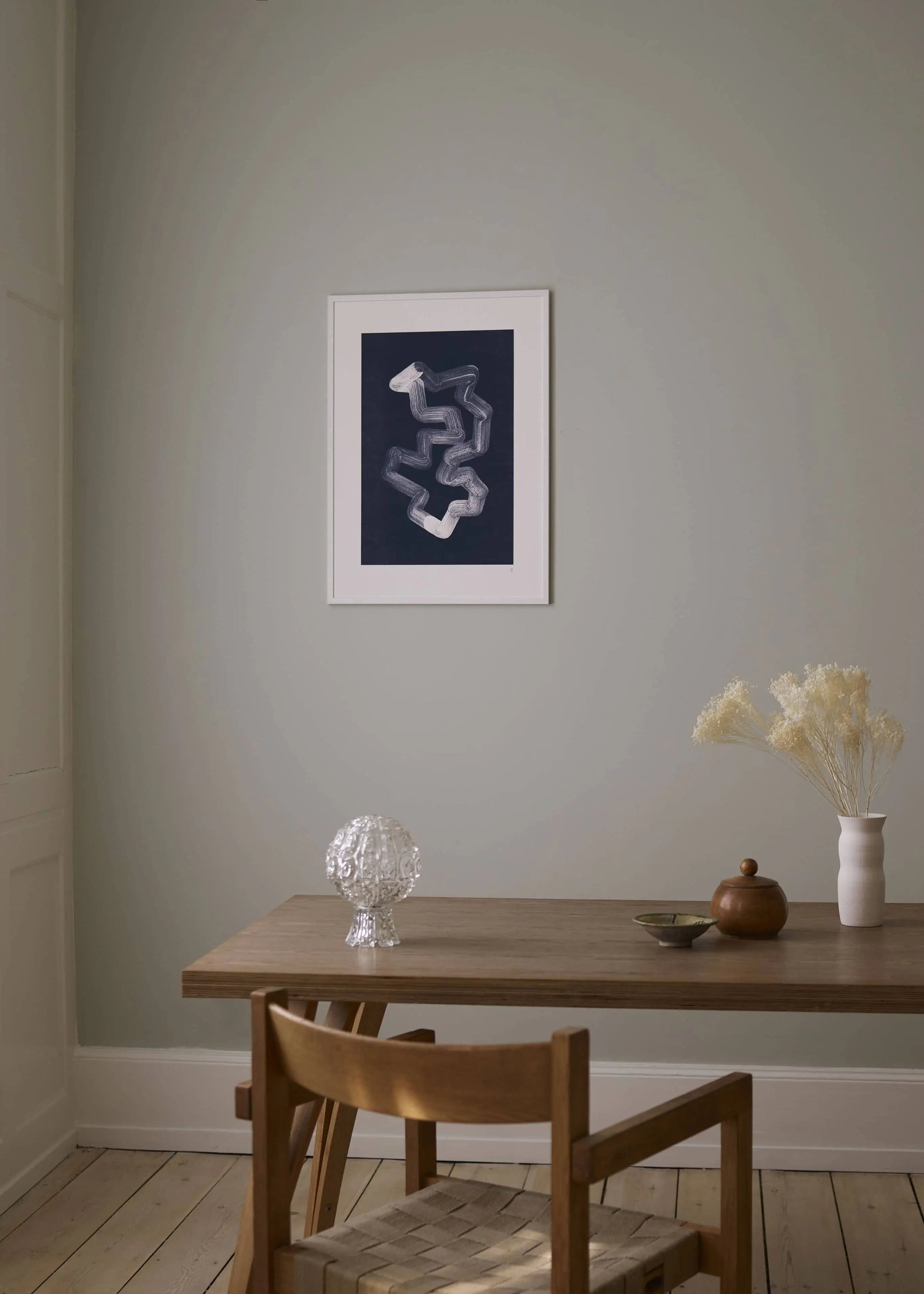 A minimalist dining area features a wooden table, two chairs, glass lamp, wooden container, bowl, and white vase with dried flowers. The Poster Club’s Jaguar 01 art print by Anastasia Krasilnikova decorates the pale gray wall.
