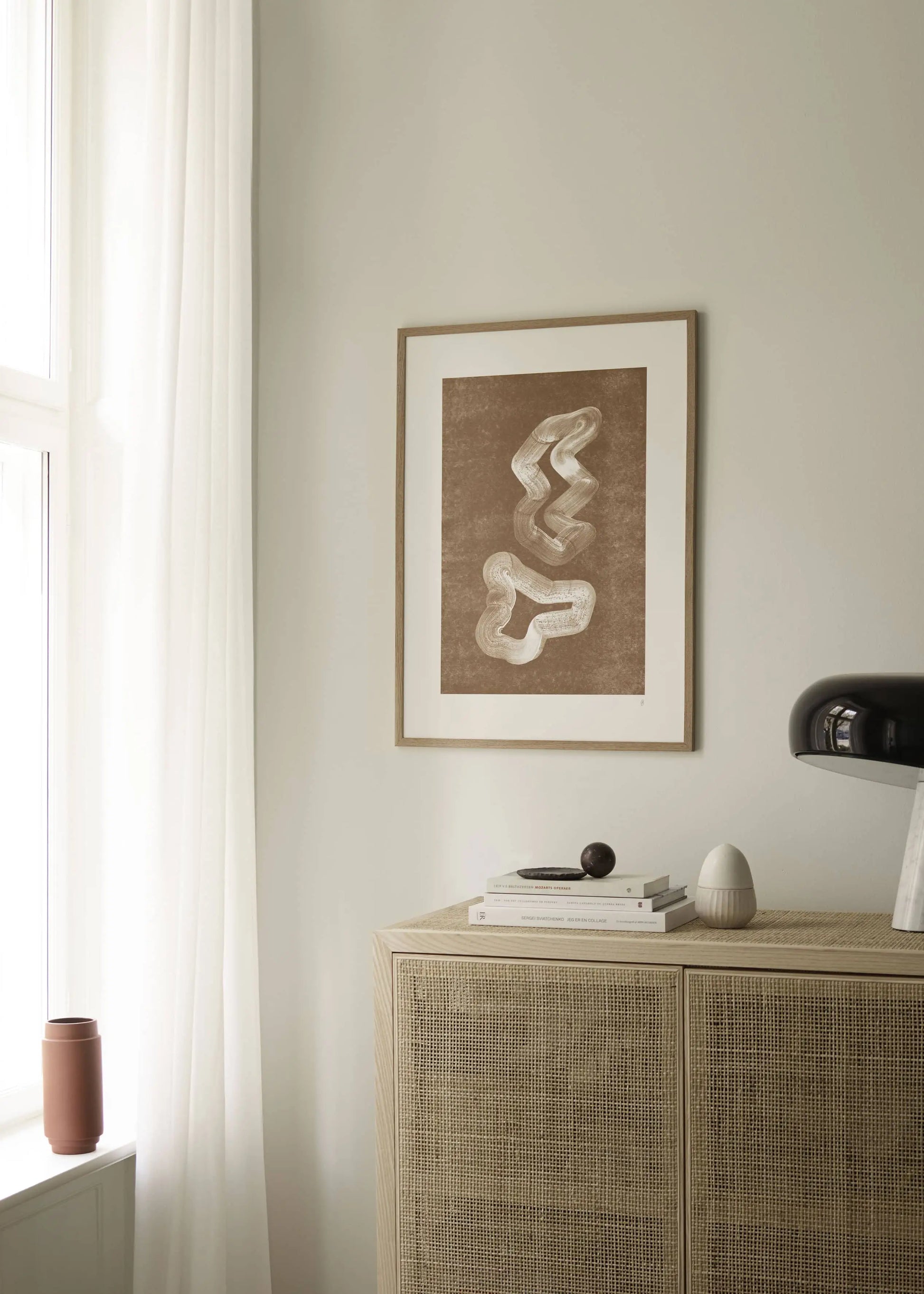 A minimalist room features light walls, sheer curtains, and The Poster Clubs Anastasia Krasilnikova - Jaguar 01 (50x70) art print in a wooden frame above a woven cabinet with books, a black lamp, and a white vase by the window.