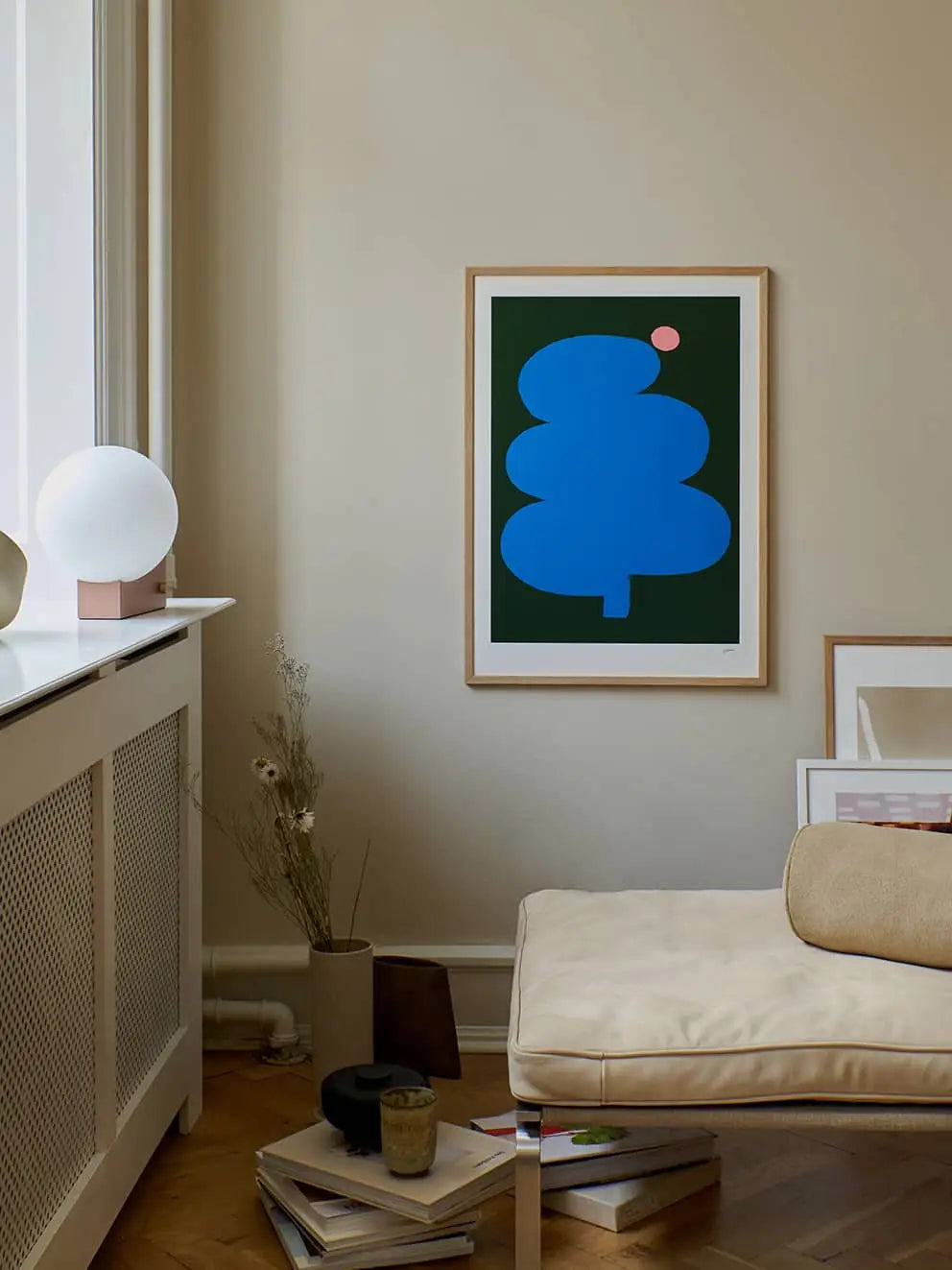 A minimal living space features a beige lounge chair, white cabinet with lamp, books and dried flowers, and The Poster Club Wensi Zhai - Fat Tree Pink Moon (50x70) giclée print of abstract blue trees on the wall.