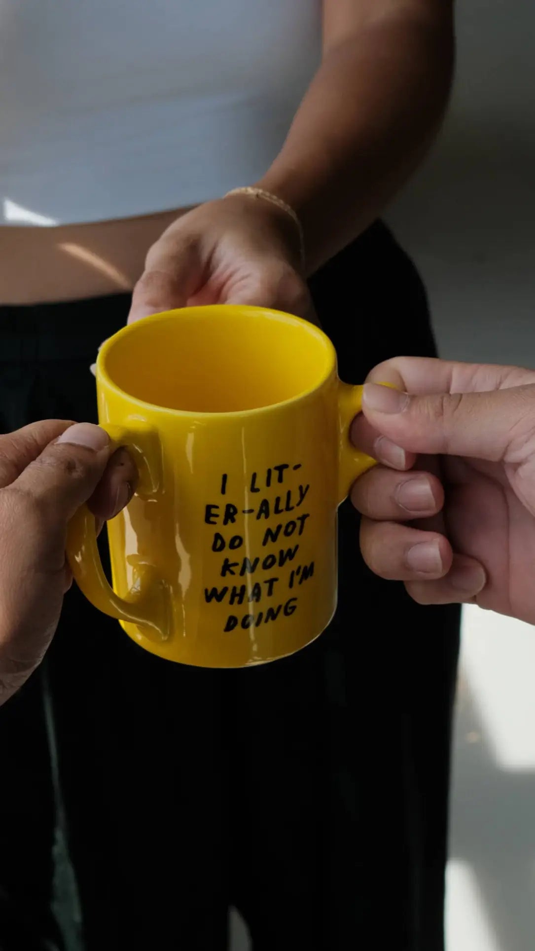 Two hands hold the Third Drawer Down Adam JK - I Literally Do Not Know Mug, a bright yellow mug with black text. In the background, someone wearing a white top and black pants is standing.