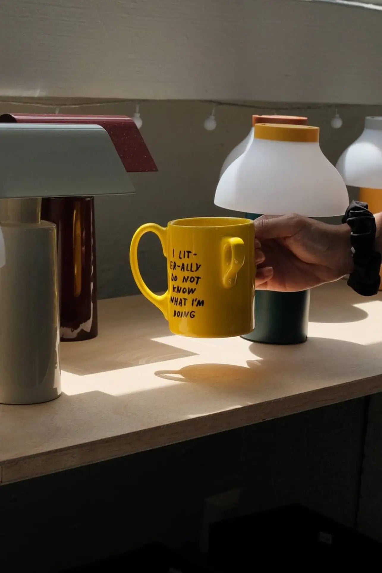 A person holds the Third Drawer Down Adam JK - I Literally Do Not Know Mug on a wooden shelf, surrounded by modern lamps in soft sunlight—a quirky Third Drawer Down piece that adds humor and style to any space.
