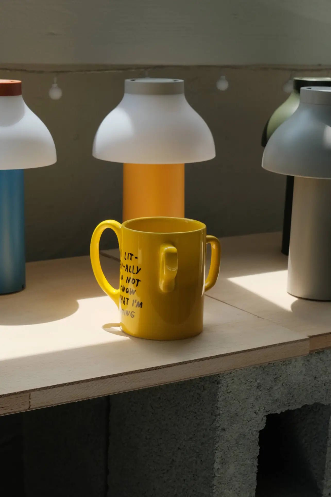 A bright yellow Third Drawer Down Adam JK - I Literally Do Not Know Mug with two handles sits on a wooden surface among colorful lamps, playfully reflecting the brand’s signature style.