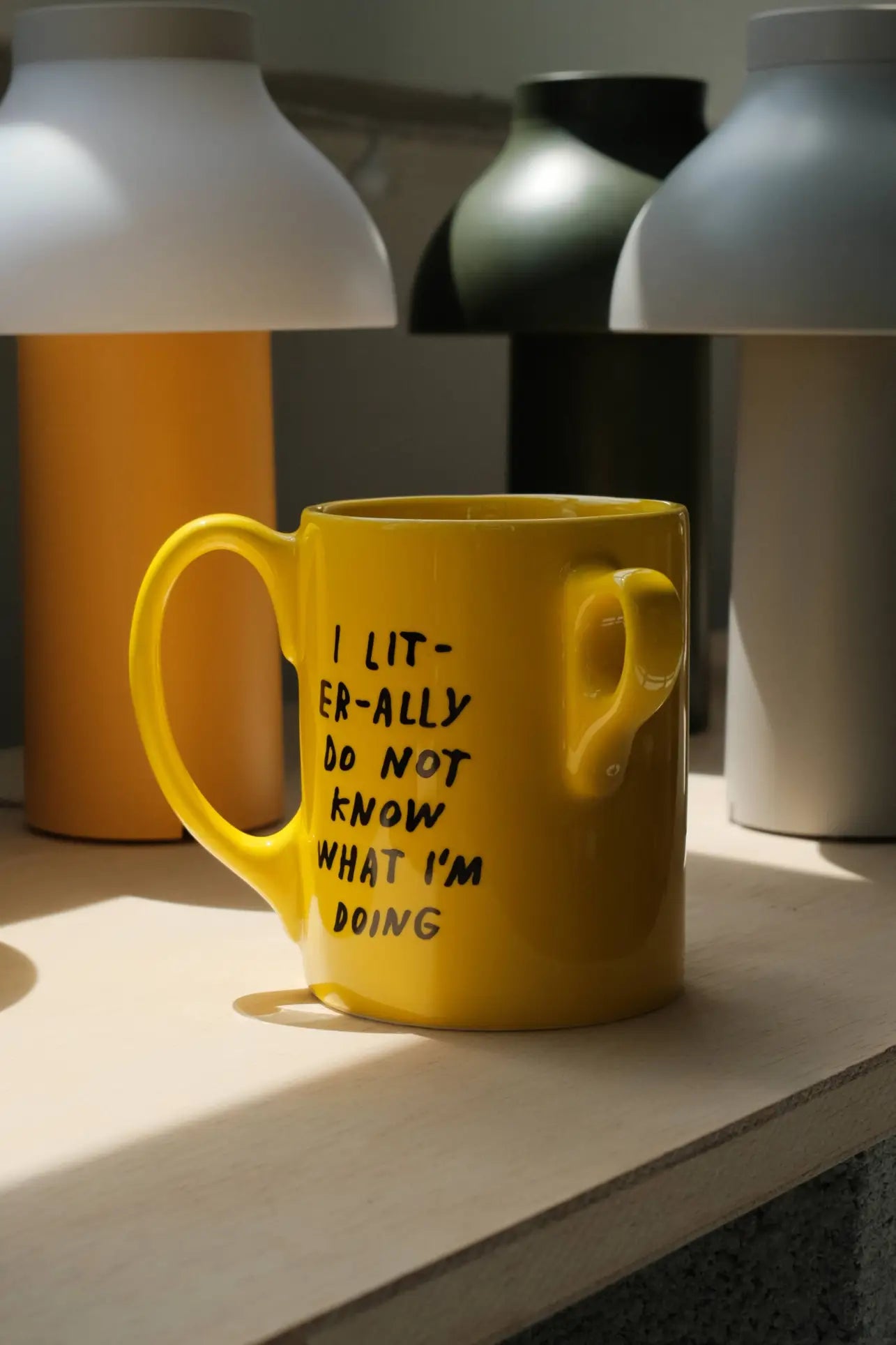 The Third Drawer Down Adam JK - I Literally Do Not Know Mug sits on a light wooden surface, featuring playful handwritten text that adds fun to your morning routine. Blurred bottles and vases appear in the background.