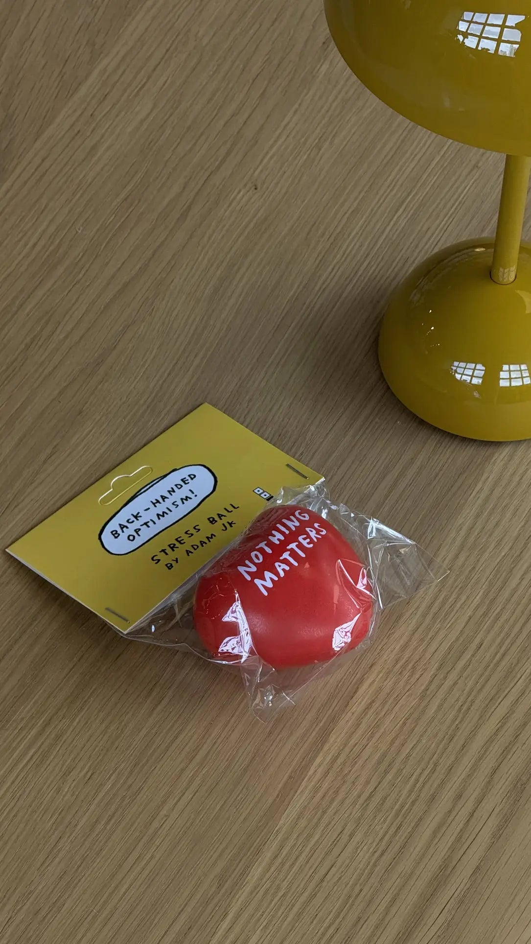A red heart-shaped stress toy from Third Drawer Down, labeled Nothing Matters, sits on a table next to a yellow lamp. The packaging reads: Adam JK - Nothing Matters Stress Toy by Third Drawer Down, offering tactile comfort and stress relief.