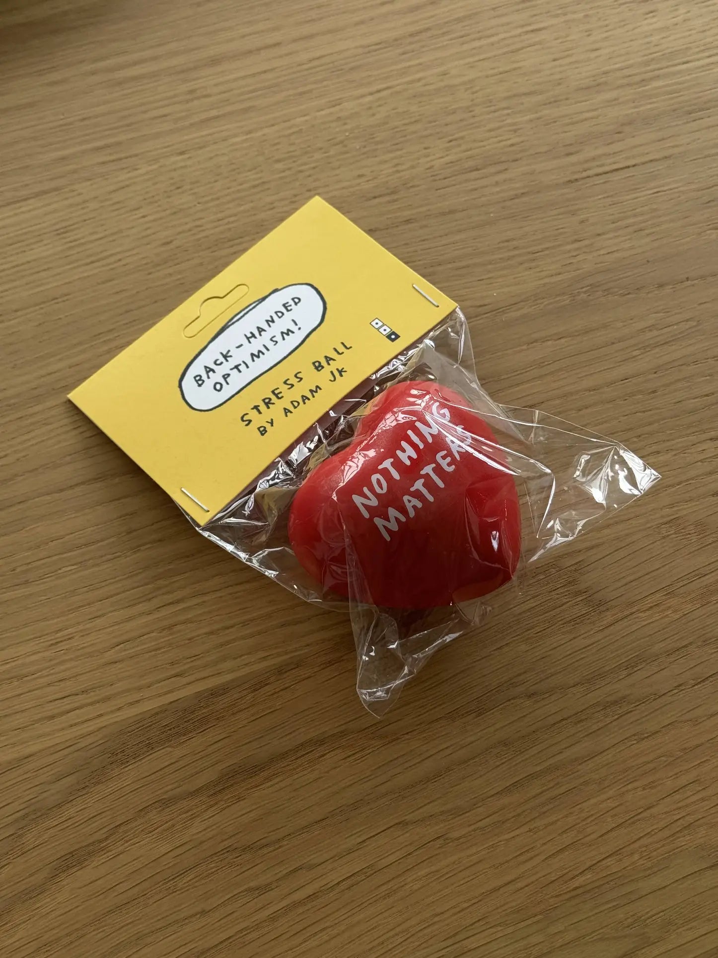 A red heart-shaped Third Drawer Down Adam JK - Nothing Matters Stress Toy in plastic packaging, labeled NOTHING MATTERS, sits on wood. A yellow tag shows its by Third Drawer Down—ideal for stress relief and tactile comfort.