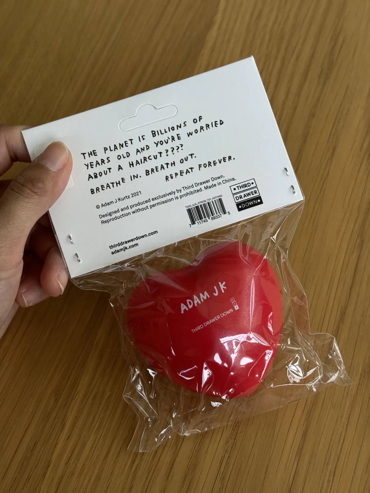 A hand holds the Third Drawer Down Adam JK - Nothing Matters Stress Toy, a red heart-shaped squishy from Third Drawer Down. The label says: The planet is billions of years old and youre worried about an email? Breathe for stress relief.