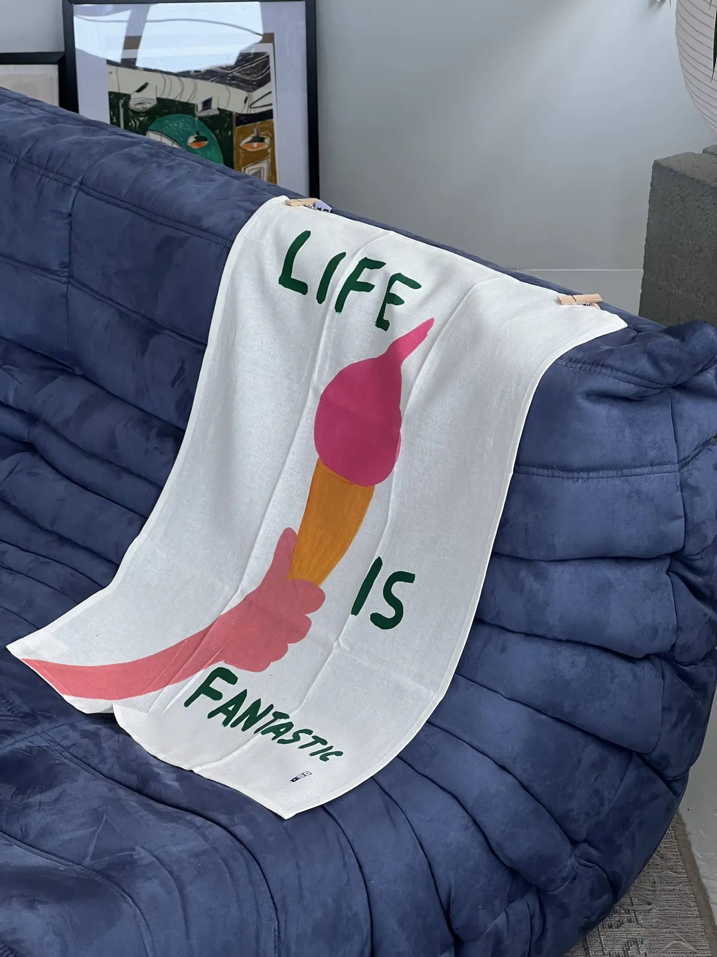 A Third Drawer Down David Shrigley - Life Is Fantastic Tea Towel with a vibrant paintbrush design and bold text hangs over a blue tufted sofa, with framed art and a gray side table in the background.