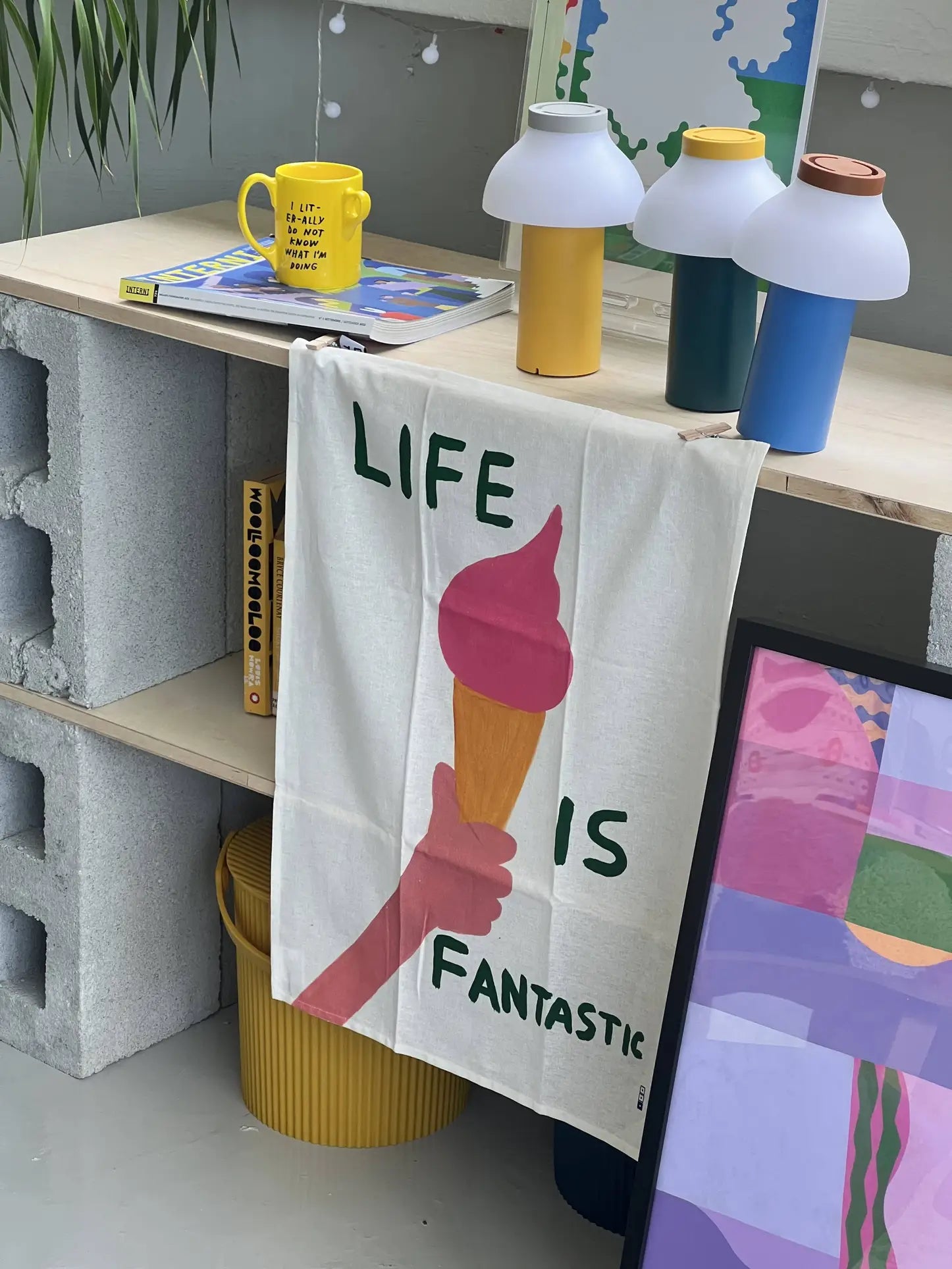 The Third Drawer Down David Shrigley - Life Is Fantastic Tea Towel, featuring a hand holding a pink ice cream and the phrase LIFE IS FANTASTIC, hangs from a wooden shelf amid colorful decor, books, lamps, and a yellow mug.