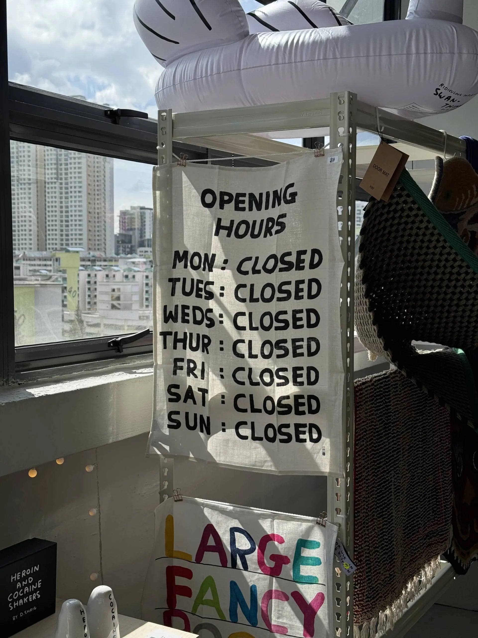 The Third Drawer Down David Shrigley - Opening Hours Tea Towel by Third Drawer Down hangs on a shelf in a sunlit store, each day marked CLOSED, with city buildings seen through the window behind.