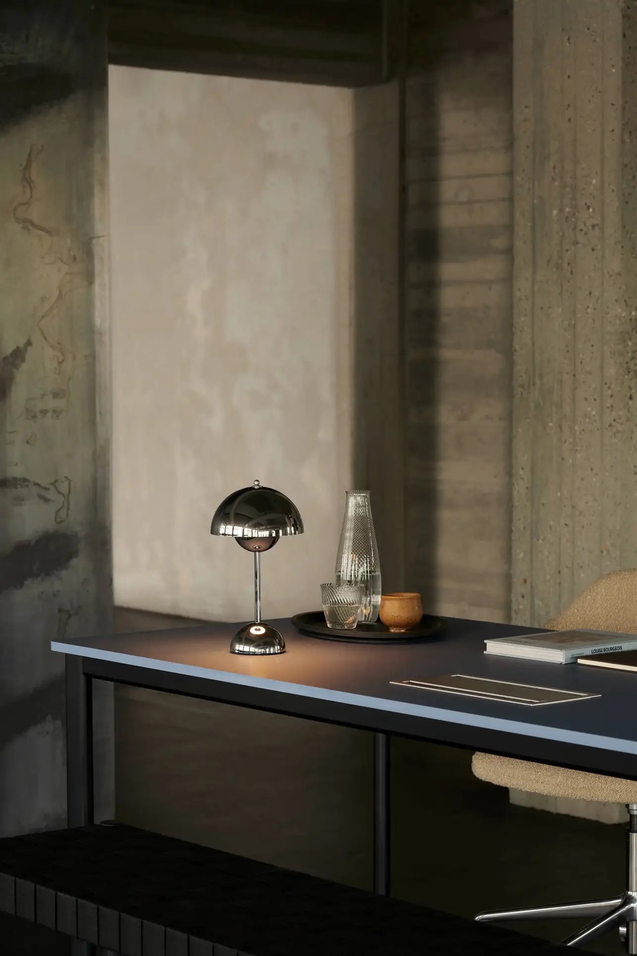 A modern desk features the &Tradition Flowerpot VP9 Portable Table Lamp, a USB charging cable, glass carafe, cup, notebook, and closed laptop set against concrete and textured walls in a minimalist office space.