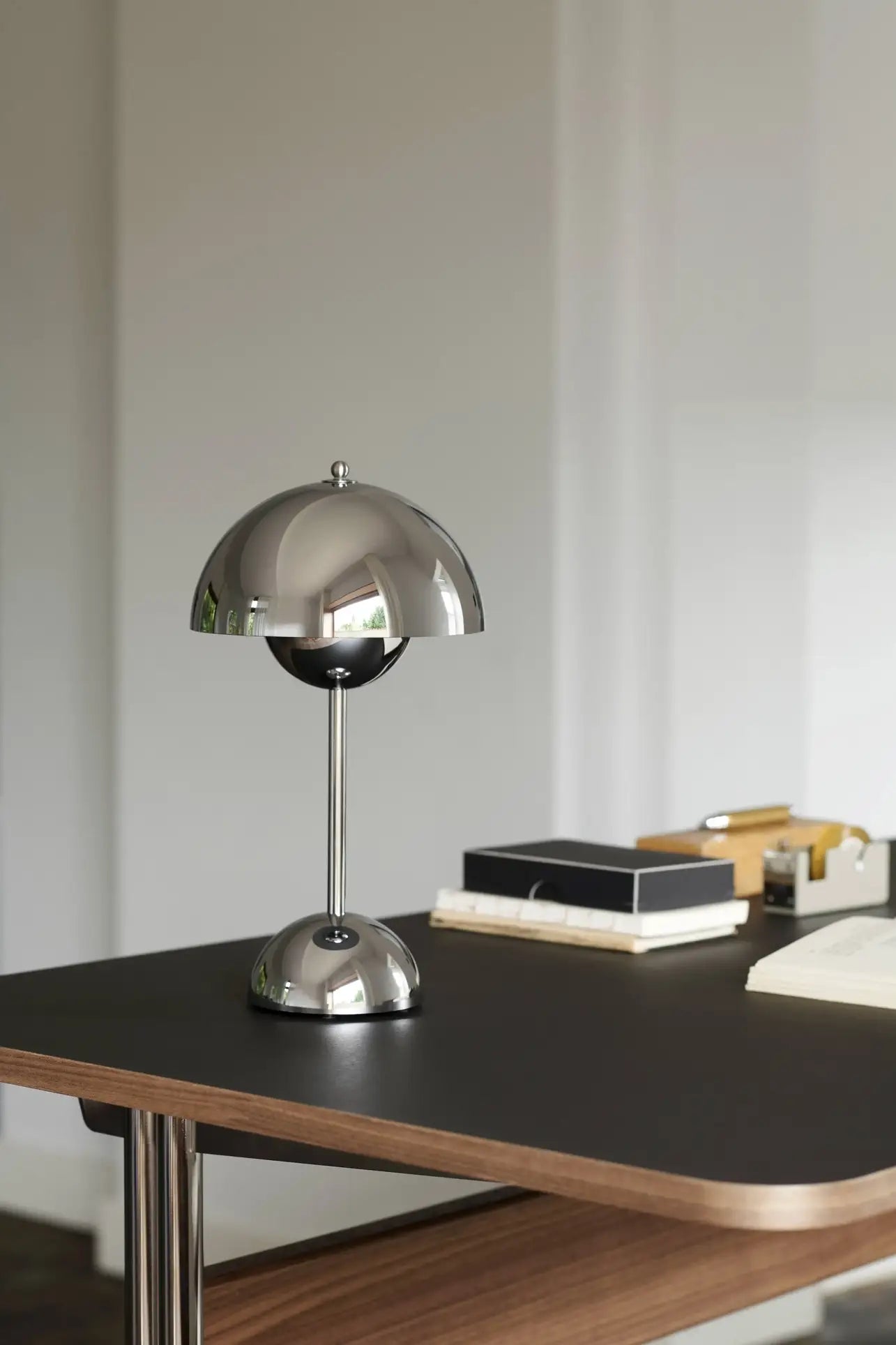 The &Tradition Flowerpot VP9 Portable Table Lamp by &Tradition, featuring a metallic dome shade, sits on a black desk with books, a notepad, tape dispenser, and USB charging cable against a neutral wall.