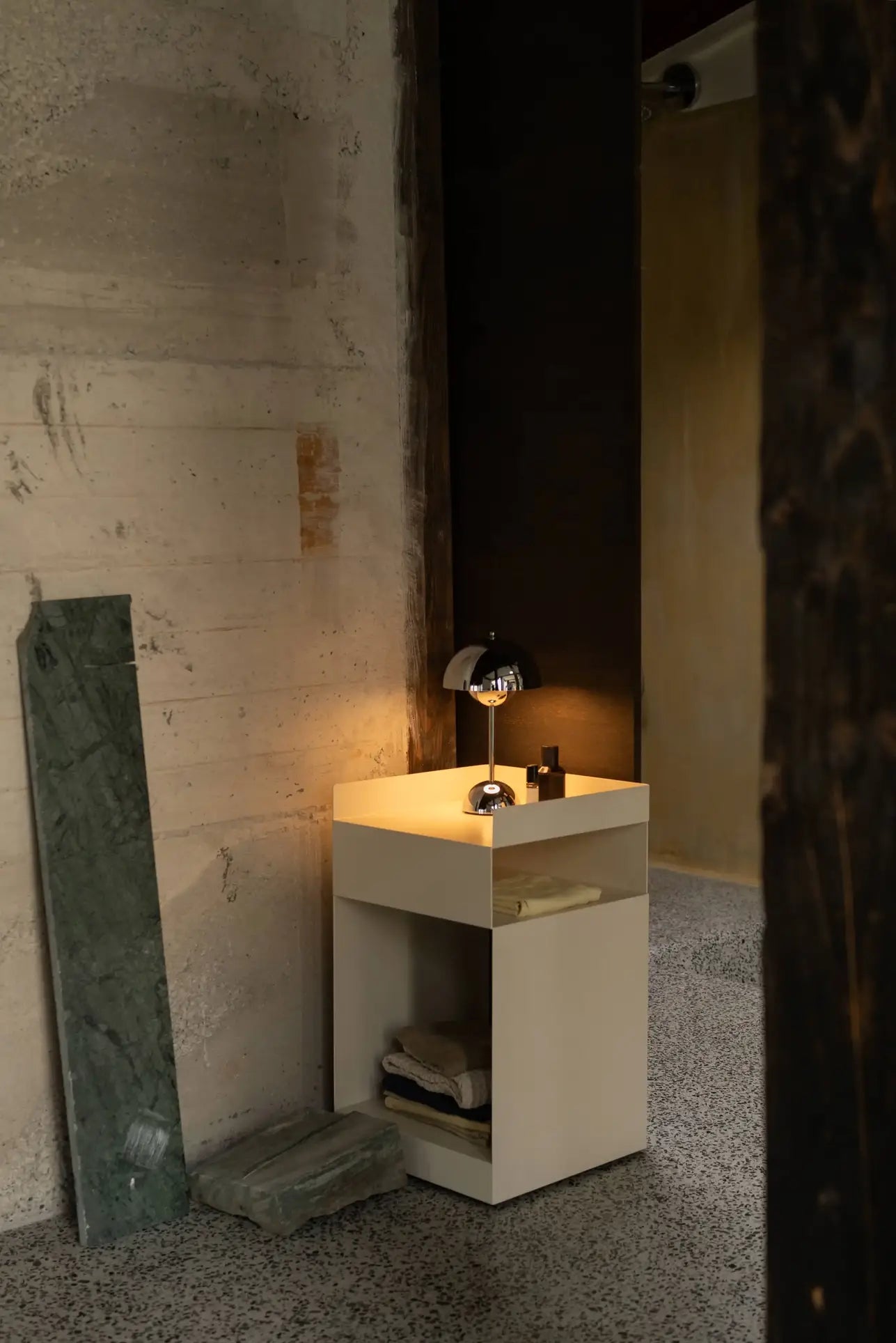 A modern, minimalist nightstand features the &Tradition Flowerpot VP9 Portable Table Lamp by &Tradition, with a few objects on top. Set against a textured green marble wall, folded towels on the lower shelf, and a discreet USB charging cable for added convenience.