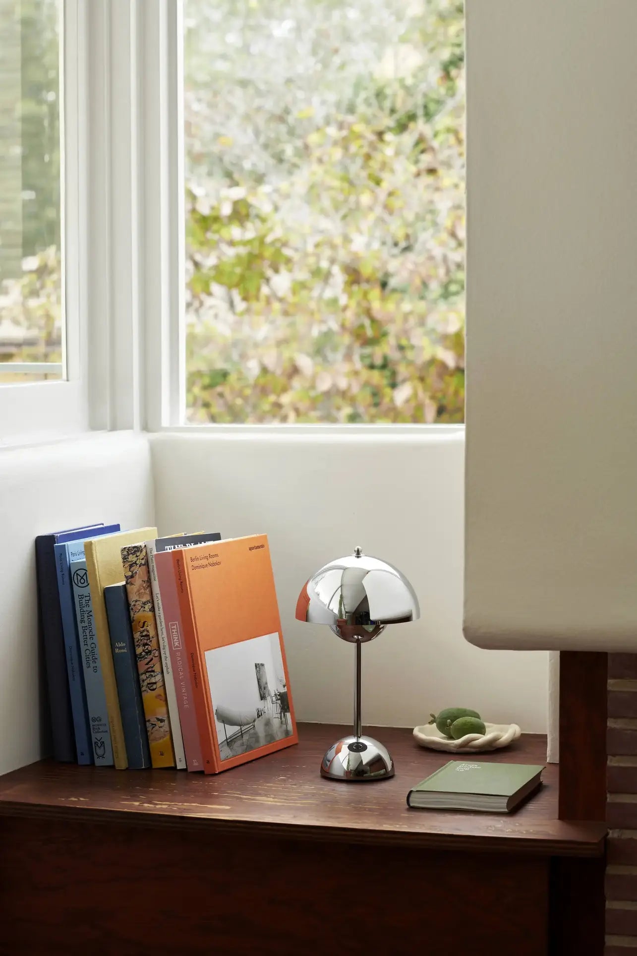 A cozy reading nook with upright books, a notebook, and a plate of green fruits rests on a wooden surface beside the window, illuminated by the &Tradition Flowerpot VP9 Portable Table Lamp, with trees visible outside.