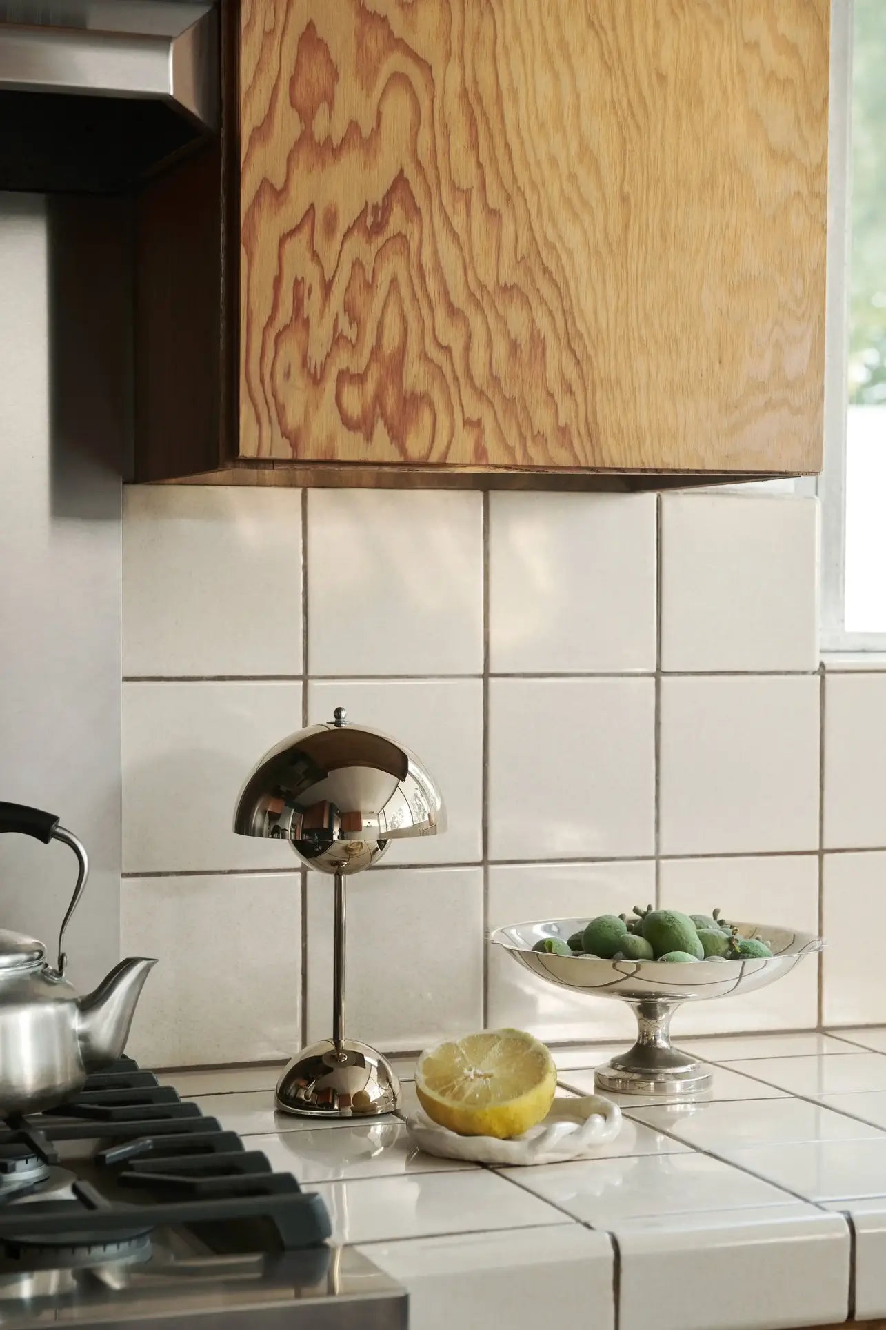 A modern kitchen counter with a stainless steel kettle, a halved lemon, green fruit in a glass dish, and the &Tradition Flowerpot VP9 Portable Table Lamp by &Tradition, all set against white tiled walls and light wood cabinetry.