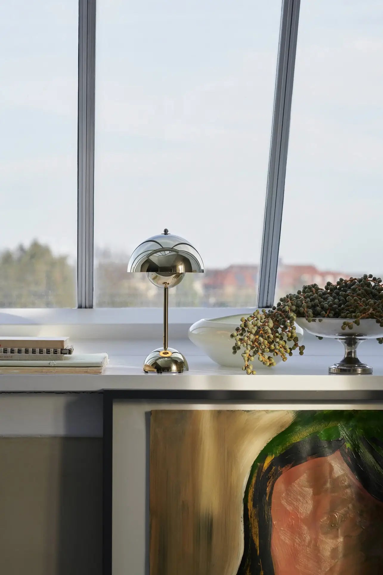 The &Tradition Flowerpot VP9 Portable Table Lamp with its metallic dome shade sits on a windowsill beside books, a white bowl, and greenery. A USB cable is nearby, and large windows with a blurred outdoor view are visible.