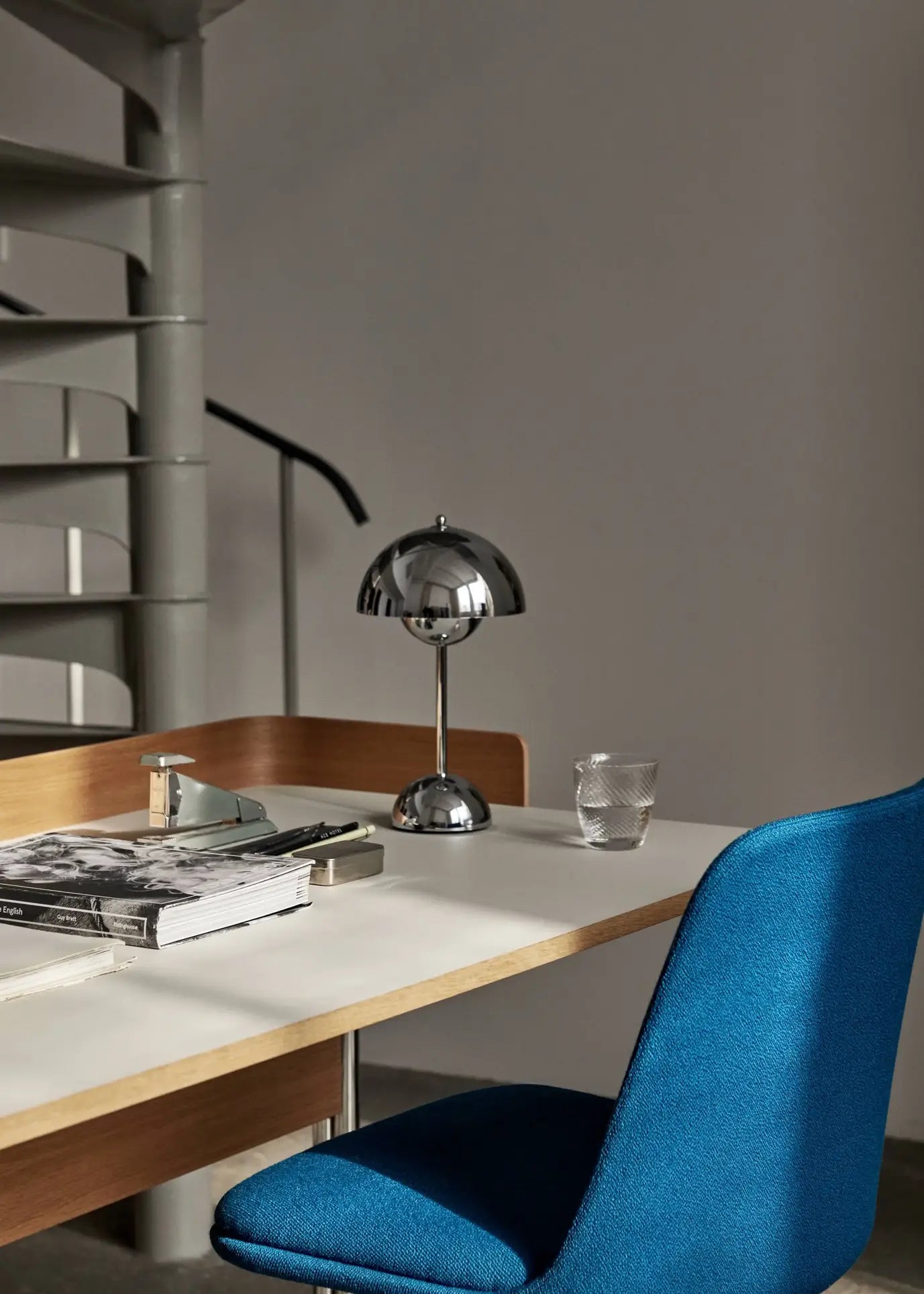 A modern workspace features a white desk, blue upholstered chair, USB cable, stacked books, glasses, a glass tumbler, and the &Tradition Flowerpot VP9 Portable Table Lamp by &Tradition. A spiral staircase is visible in the background.