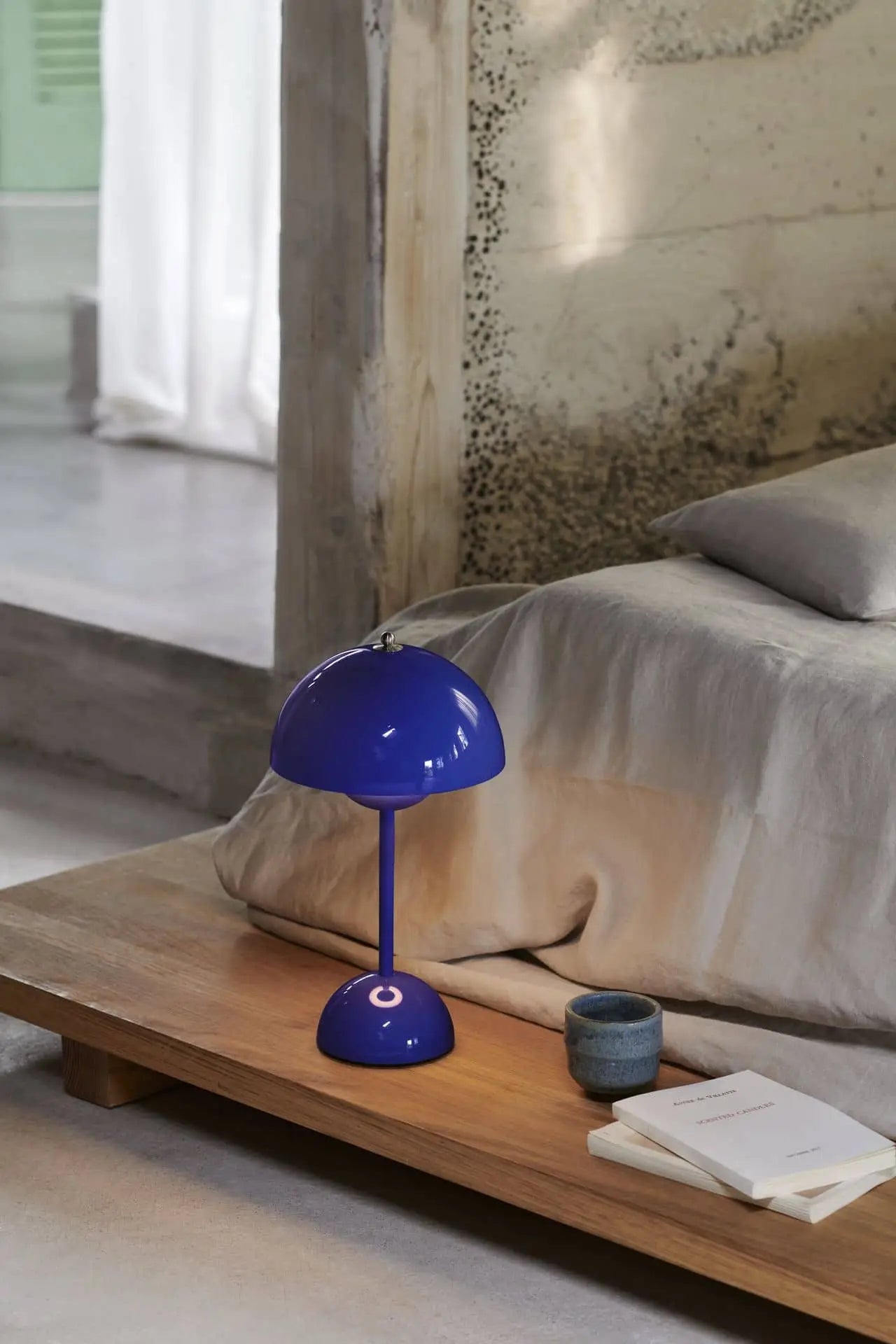 A modern bedroom showcases a low wooden bedside table with the &Tradition Flowerpot VP9 Portable Table Lamp, a ceramic cup, two books, and a USB cable. The bed with light bedding sits by a textured wall beside sheer curtains filtering sunlight.