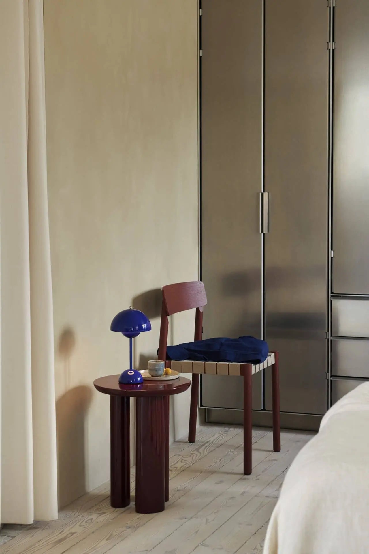 A modern bedroom corner features a small wooden table with the &Tradition Flowerpot VP9 Portable Table Lamp, ceramic decor, and a wooden chair with a blue cushion next to metallic wardrobes and cream curtains.