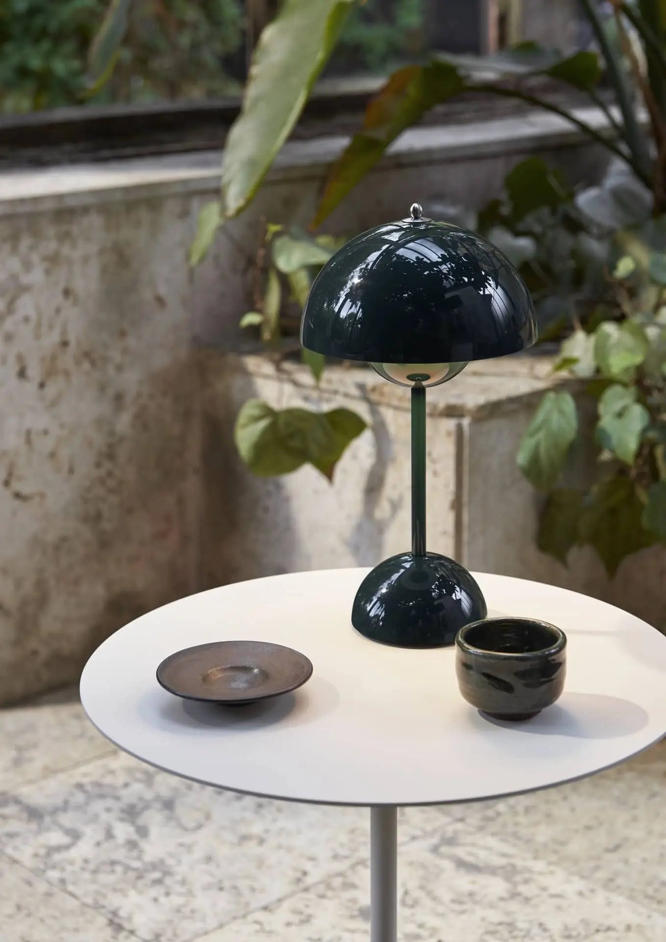 A round white table displays an &Tradition Flowerpot VP9 Portable Table Lamp in dark green, a black ceramic bowl, and a black cup. A USB charging cable is beside the lamp, with large leafy plants and a stone wall in the background.