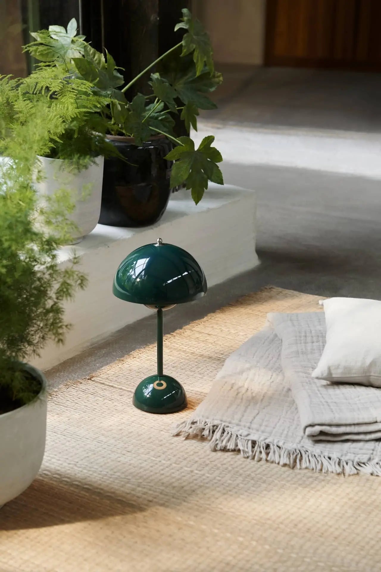 A small green &Tradition Flowerpot VP9 Portable Table Lamp sits on a woven rug beside beige blankets and white pillows, surrounded by lush plants in a cozy, sunlit room—perfect for using its handy USB charging cable.
