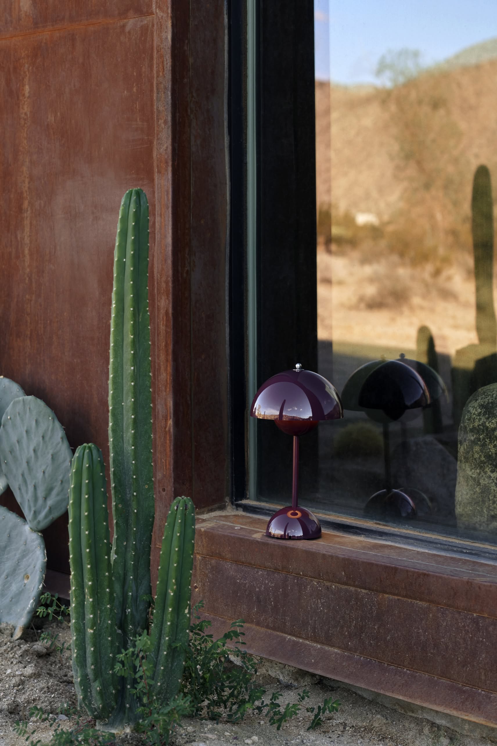 A tall green cactus stands by a rust-colored metal wall and window. On the ledge sits an &Tradition Flowerpot VP9 Portable Table Lamp in dark red, its reflection visible in the glass with a desert landscape beyond.