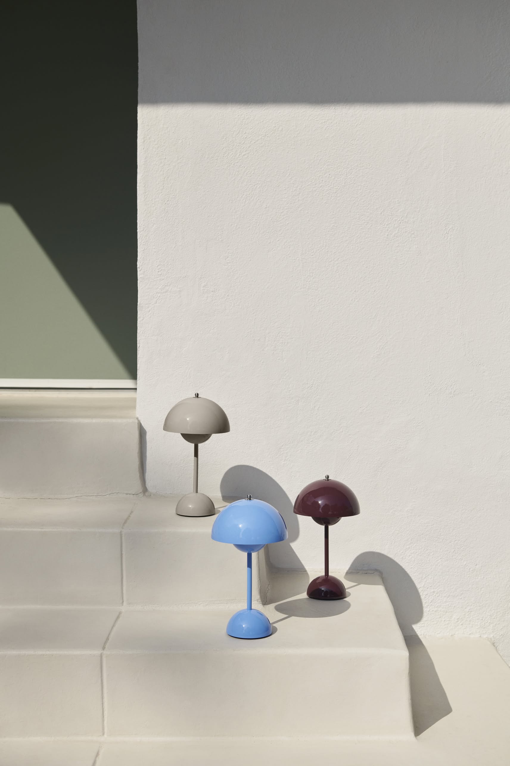 Three &Tradition Flowerpot VP9 Portable Table Lamps in light gray, light blue, and dark burgundy are displayed on pale steps against a white wall. Each lamp includes a USB charging cable for easy portability.