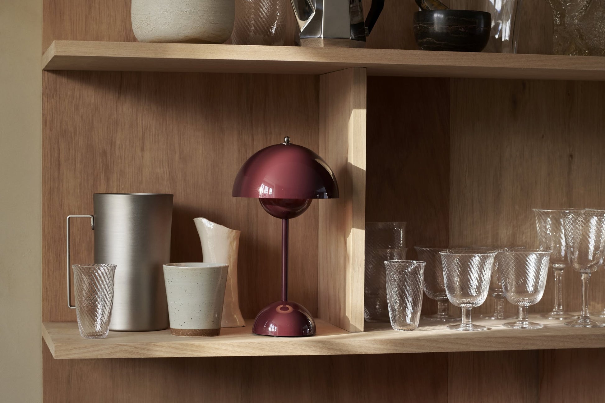 A wooden shelf displays glassware, a metallic pitcher, ceramic cups, and the &Tradition Flowerpot VP9 Portable Table Lamp with a dome-shaped shade. The lamp offers convenient USB charging for flexibility in any space.