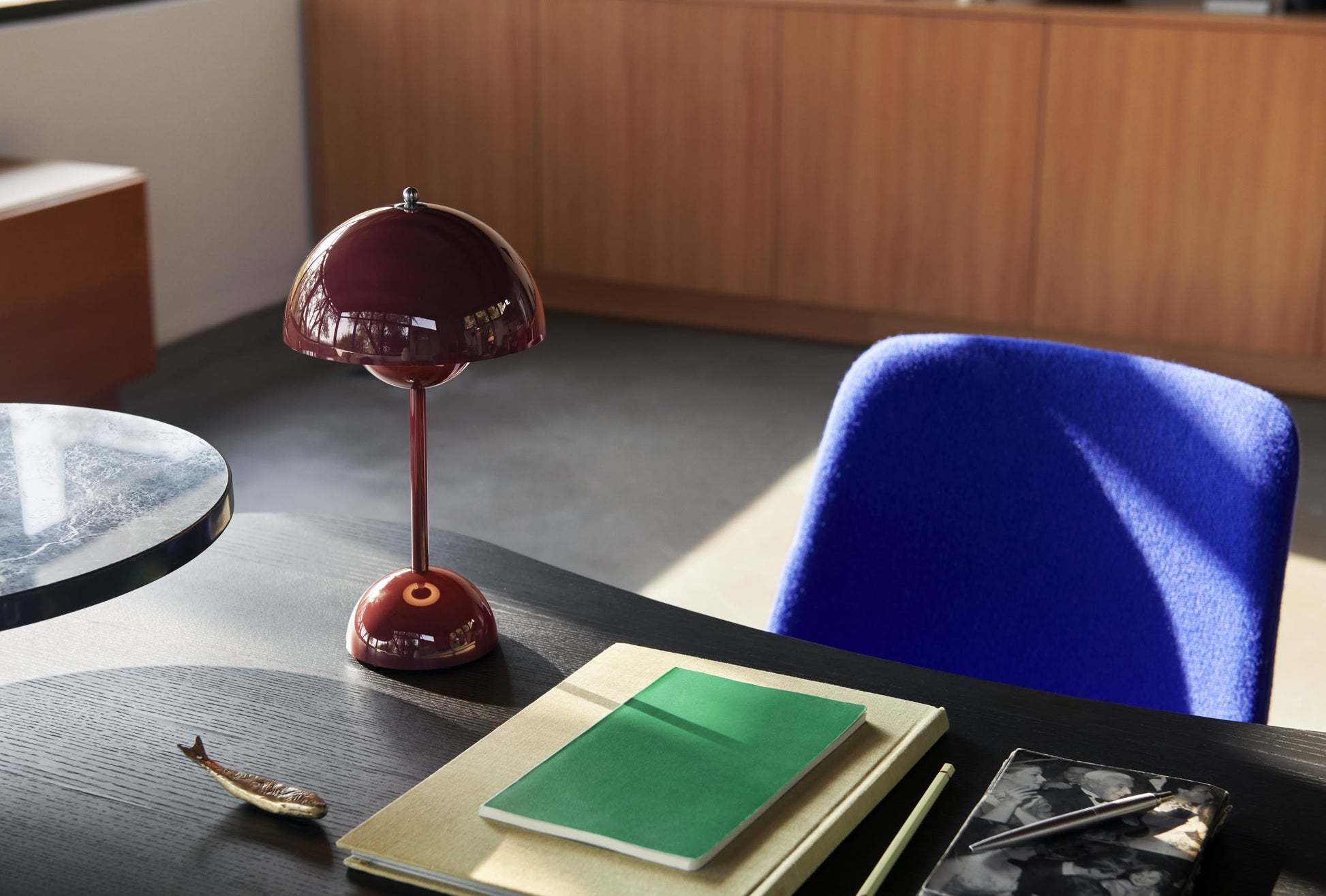 A modern desk with a blue chair features the &Tradition Flowerpot VP9 Portable Table Lamp, a green notebook, pen, closed folder, small fish-shaped object, and USB charging cable against wooden cabinets. Sunlight partially lights the scene.