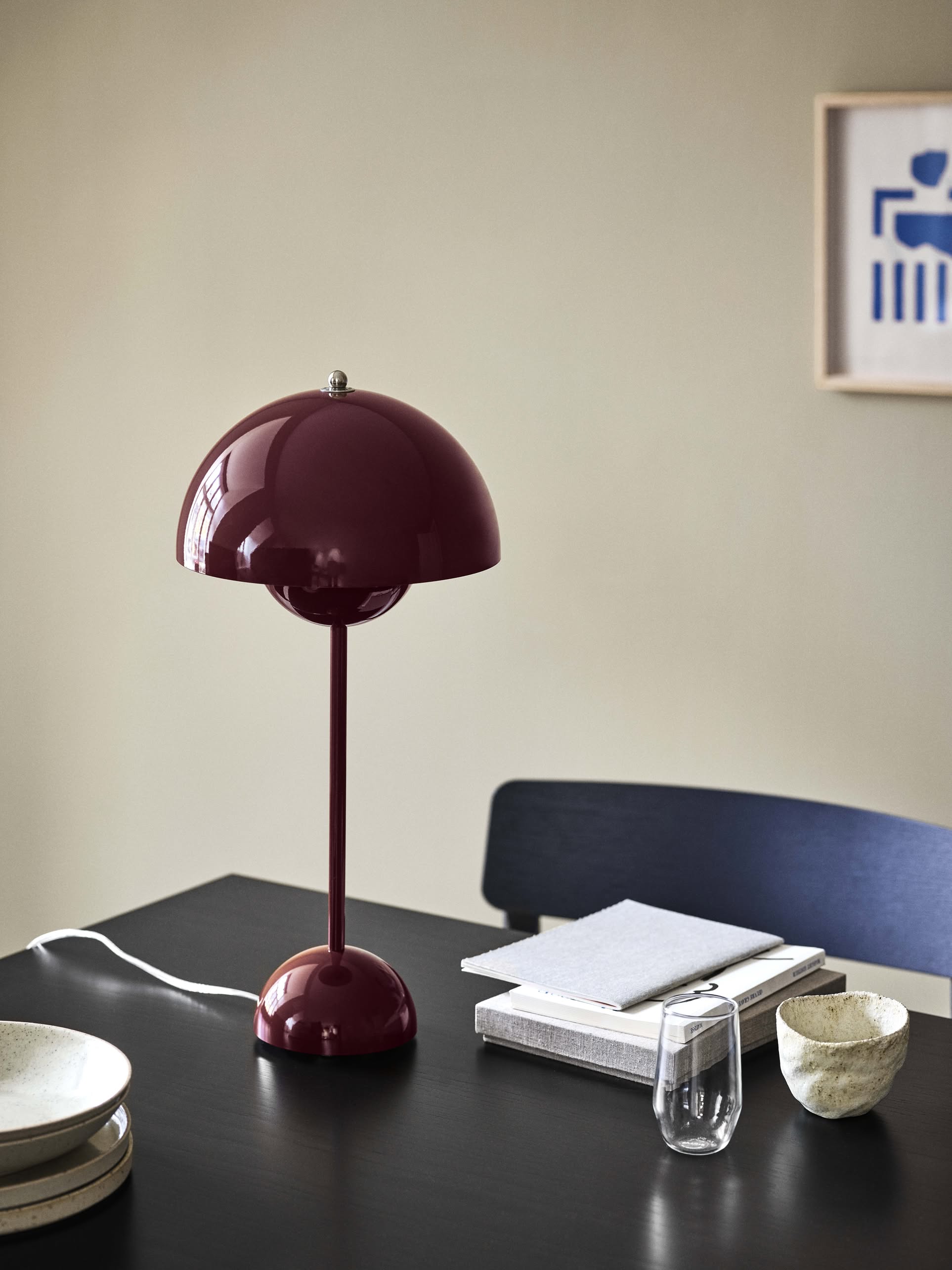 A modern workspace with a dark table featuring the &Tradition Flowerpot VP9 Portable Table Lamp in glossy maroon, stacked books, a notepad, glass of water, ceramic bowls, a dark chair, and abstract art on a beige wall.