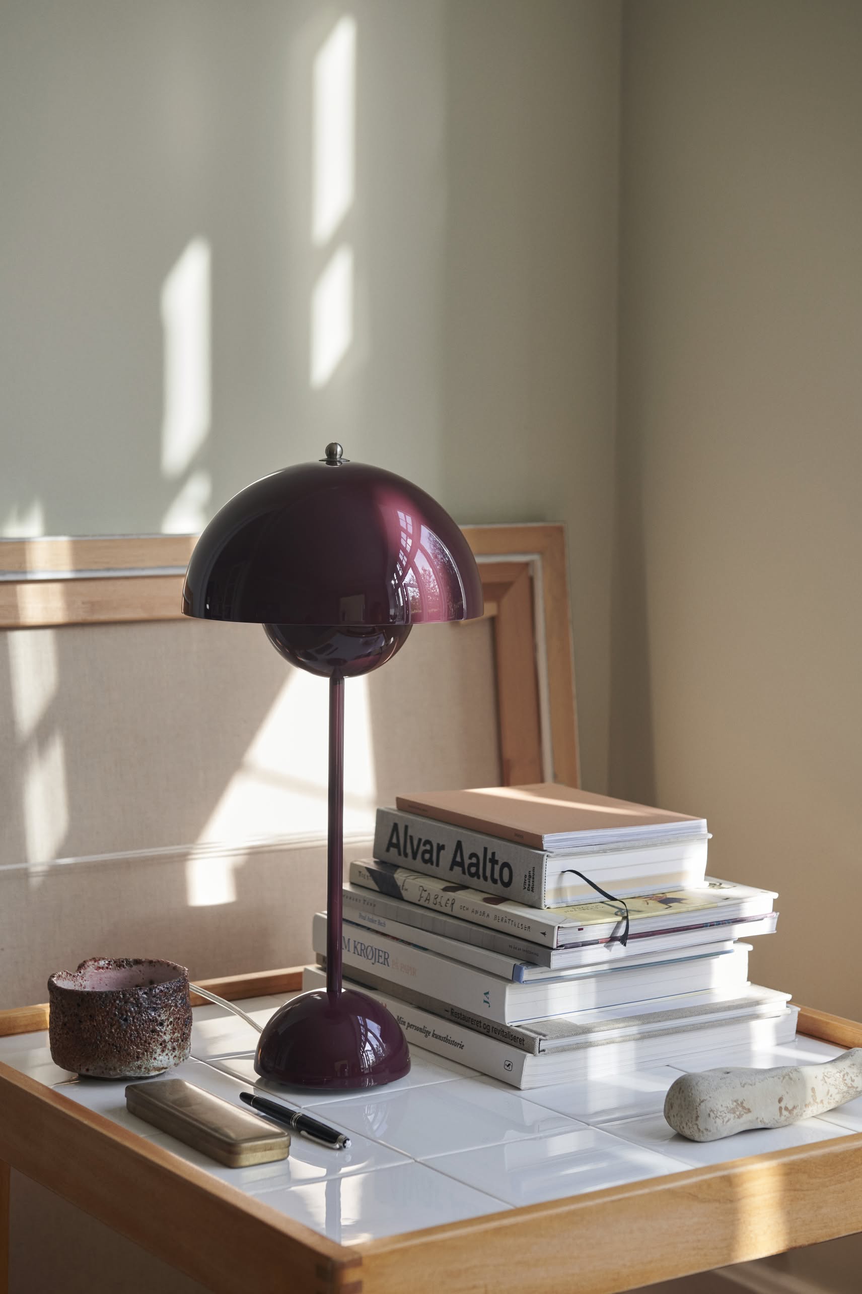 The &Tradition Flowerpot VP9 Portable Table Lamp in burgundy sits on a white surface beside design books, a candle, and a stone, with sunlight casting shadows on the beige wall behind.