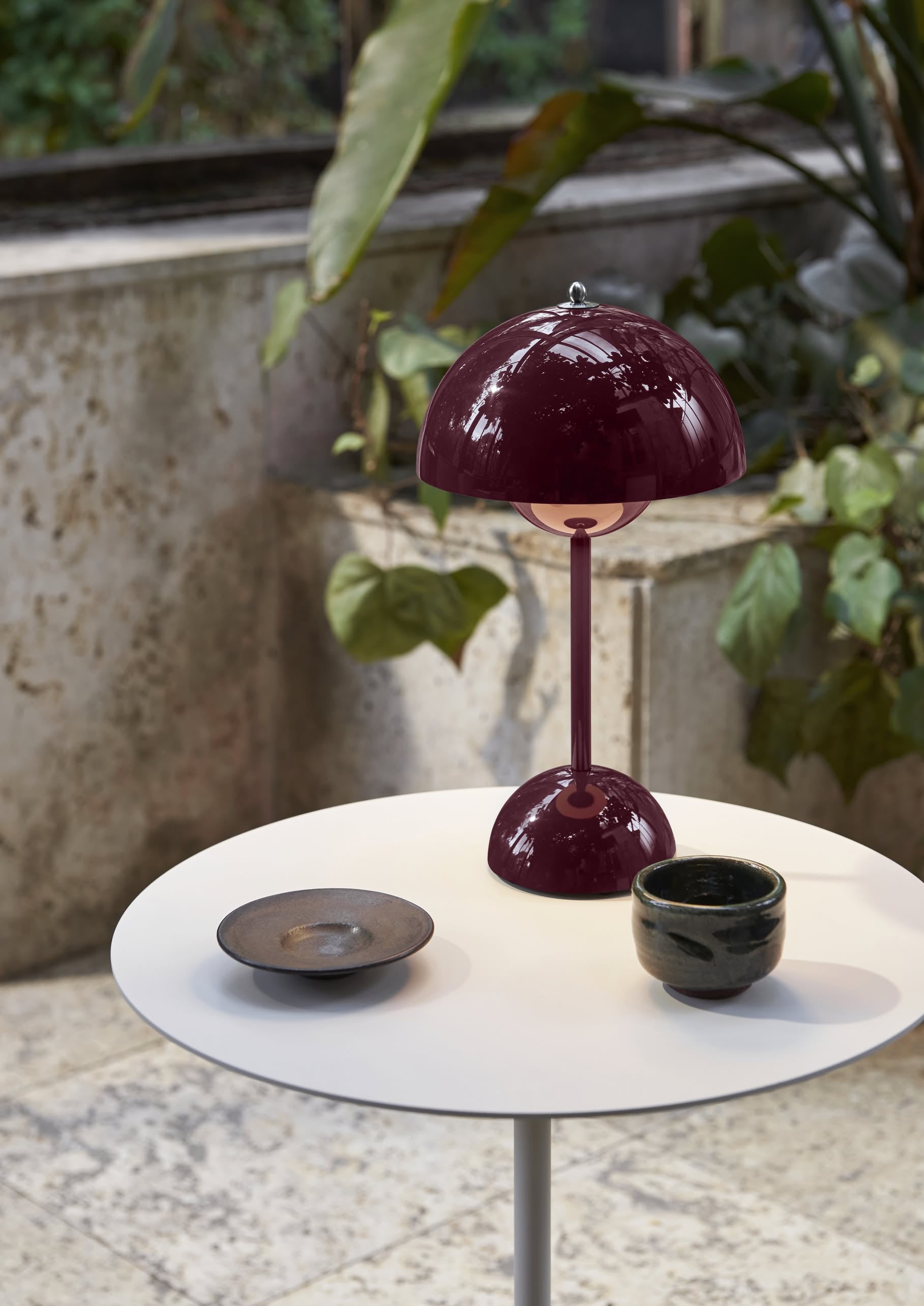 A small white round table displays an &Tradition Flowerpot VP9 Portable Table Lamp in dark red, a ceramic cup, and a dark plate with a USB charging cable nearby. Green plants and a textured stone wall form the backdrop.