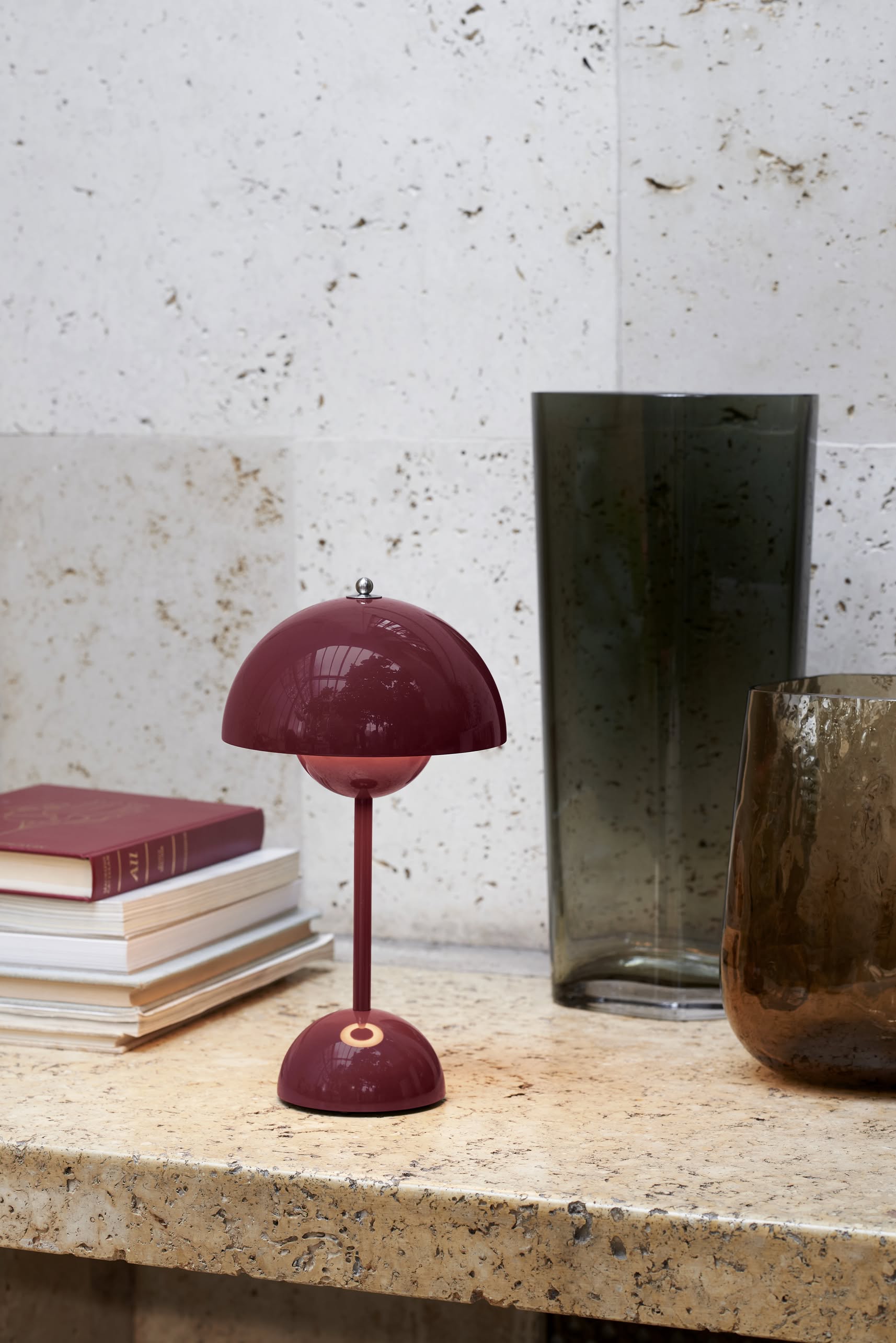 The &Tradition Flowerpot VP9 Portable Table Lamp by &Tradition, featuring a dome shade in burgundy, sits on stone beside stacked books and glass vases. This portable lamp comes with a USB charging cable and is set against a pale textured stone wall.