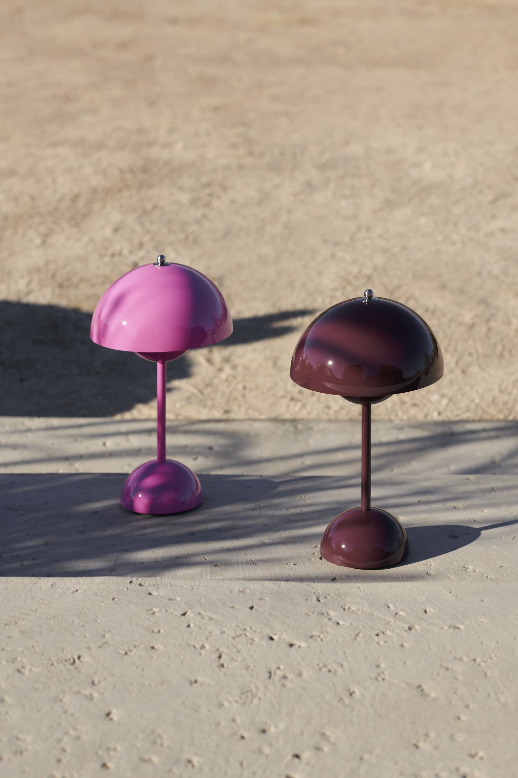 Two modern, dome-shaped &Tradition Flowerpot VP9 Portable Table Lamps in pink and dark purple stand outdoors on a light surface with sandy ground in the background. Each lamp includes a USB charging cable for easy portability.