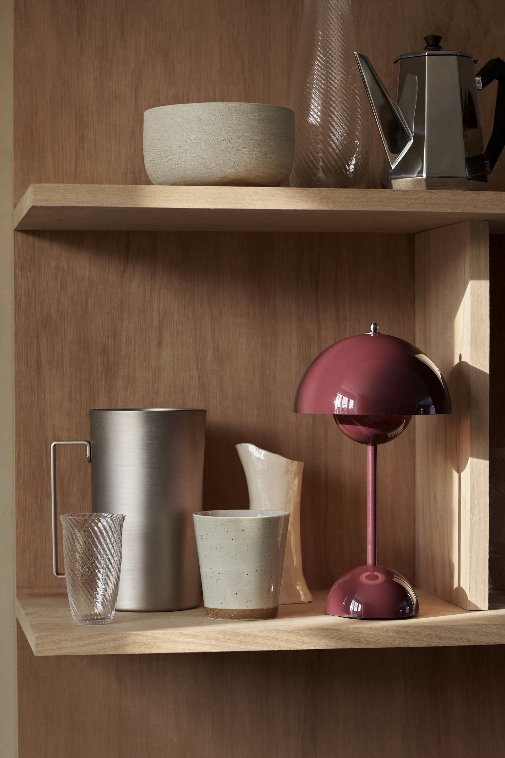 A wooden shelf displays ceramic bowls, a glass tumbler, a clear vase, a steel pitcher, a cream jug, and an &Tradition Flowerpot VP9 Portable Table Lamp in burgundy. A metal teapot sits on the top shelf.