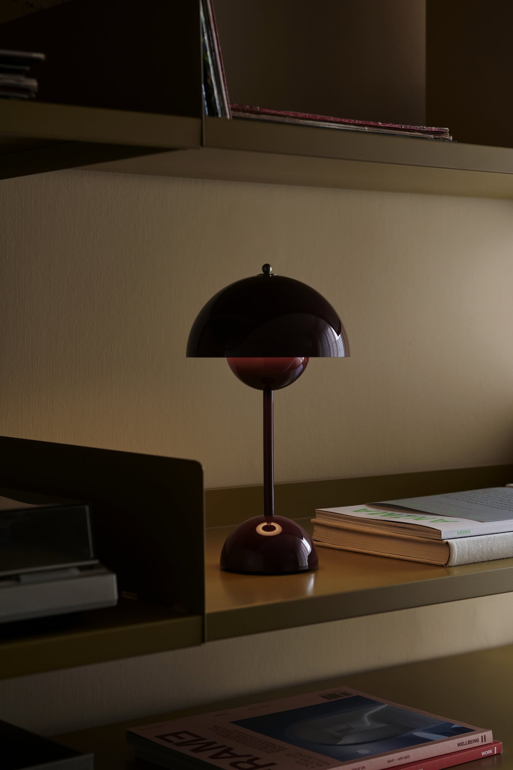 The &Tradition Flowerpot VP9 Portable Table Lamp in dark burgundy sits on a shelf beside a USB charging cable, a closed book, and magazines, adding soft light to the modern interior.