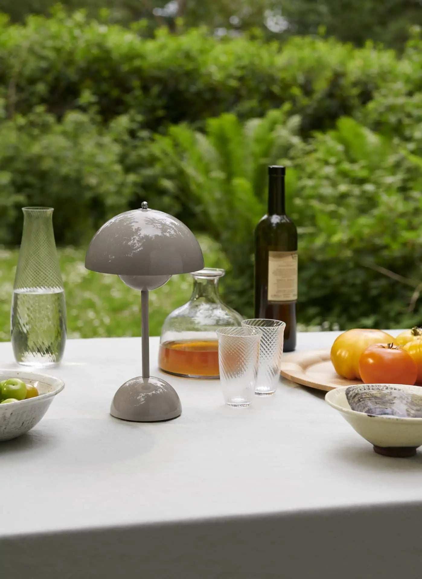 An outdoor table features the &Tradition Flowerpot VP9 Portable Table Lamp, a decanter of amber liquid, a wine bottle, glassware, bowls of green olives and fruit, and a wooden board with tomatoes, set against lush green foliage.