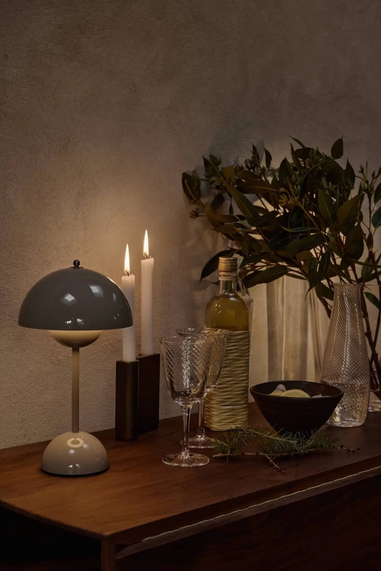 A warmly lit wooden table features the &Tradition Flowerpot VP9 Portable Table Lamp, two lit candles, wine glasses, a bottle, a bowl, and a vase with leafy branches set against a textured beige wall.