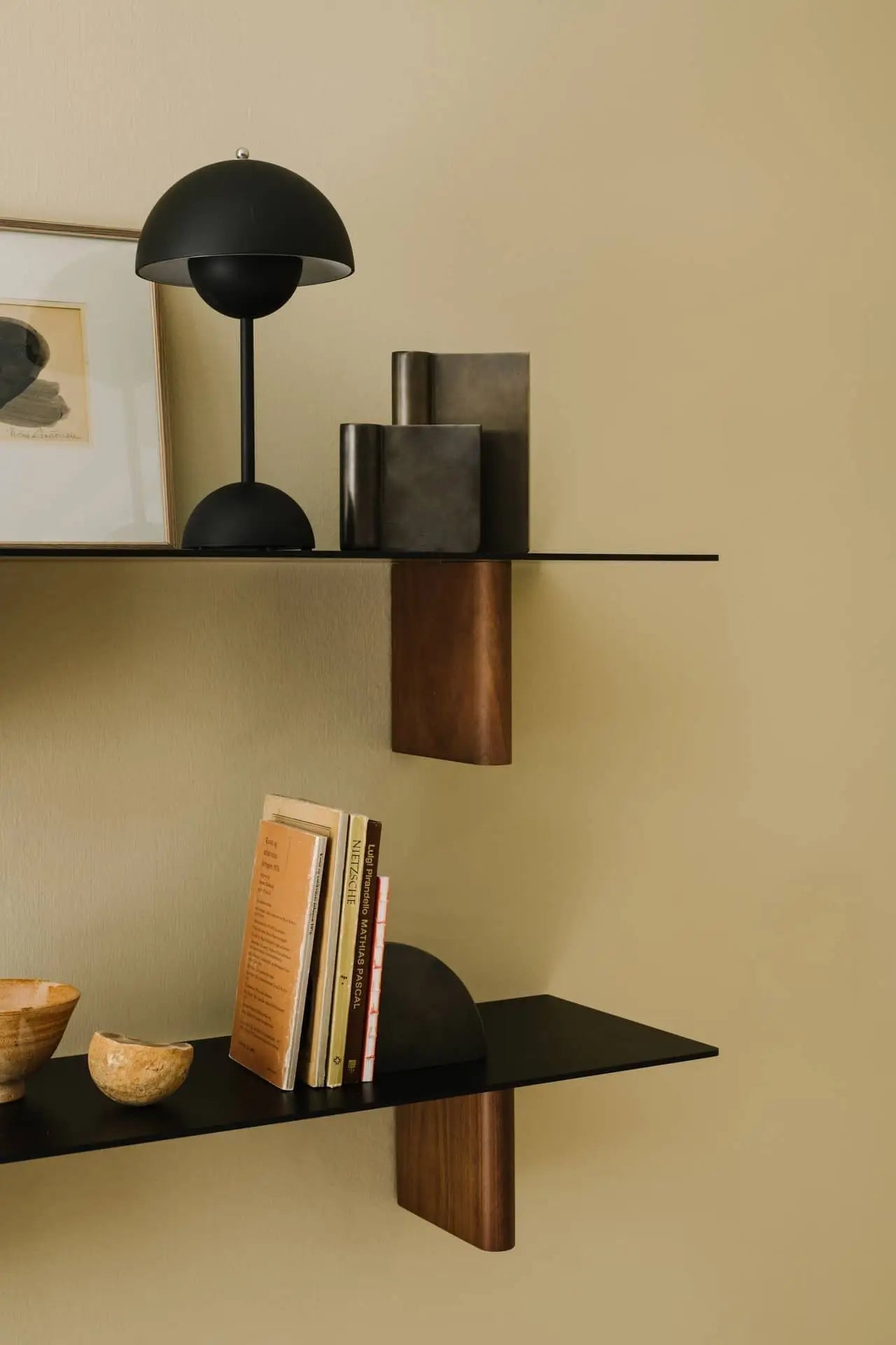 Minimalist shelves on a beige wall display books, small wooden bowls, the &Tradition Flowerpot VP9 Portable Table Lamp, decorative objects, and framed art for a clean, contemporary look.
